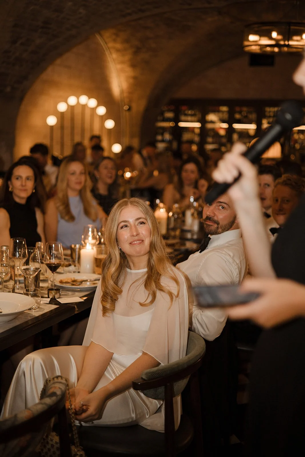 A woman with long blonde hair sitting at a dinner table during a formal event, smiling, with a man with a beard sitting beside her, looking at her. The background shows other guests, dim warm lighting, and a bar or shelf.