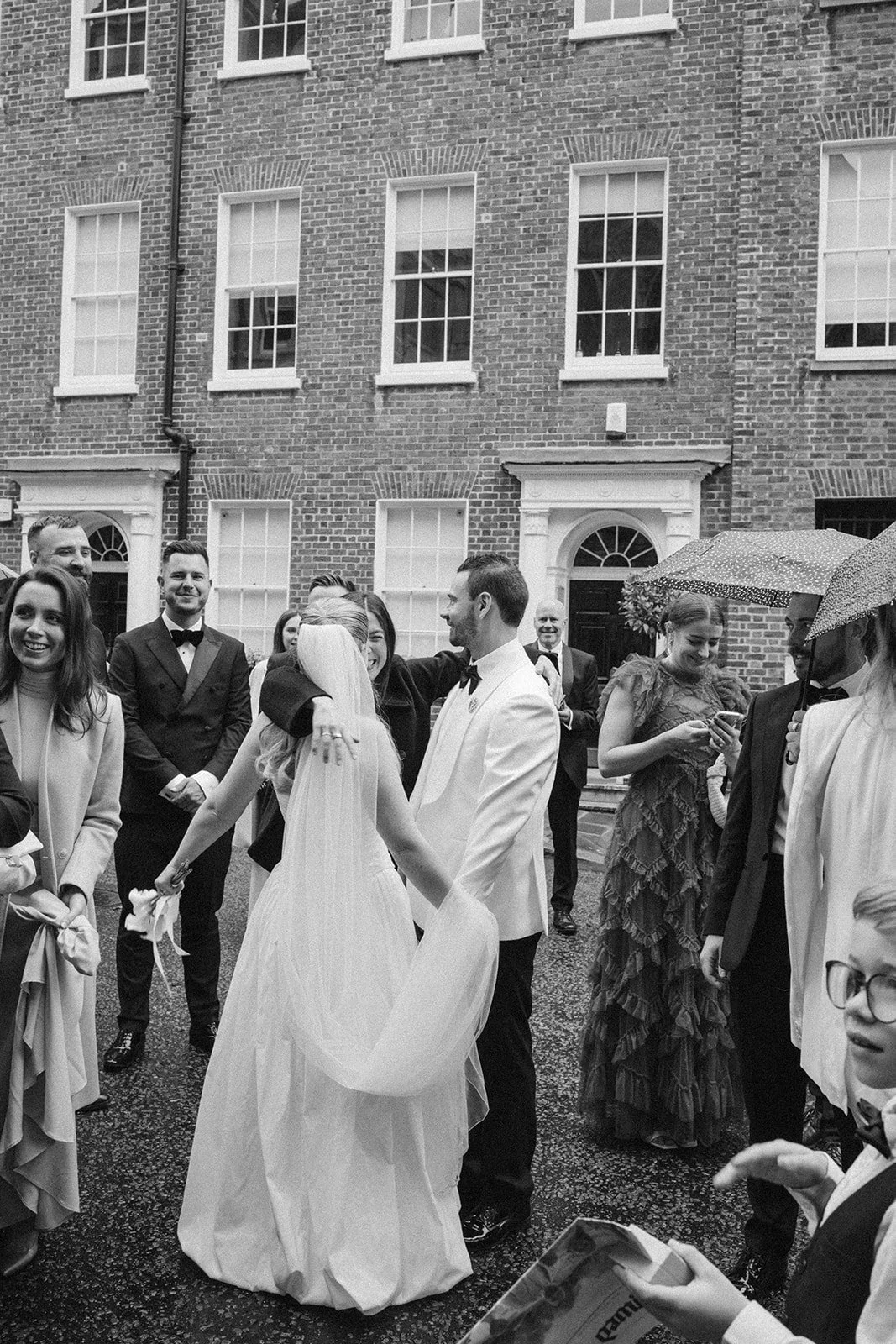 A bride and groom holding hands and smiling at each other, surrounded by wedding guests on a rainy day outside a brick building, some guests holding umbrellas.