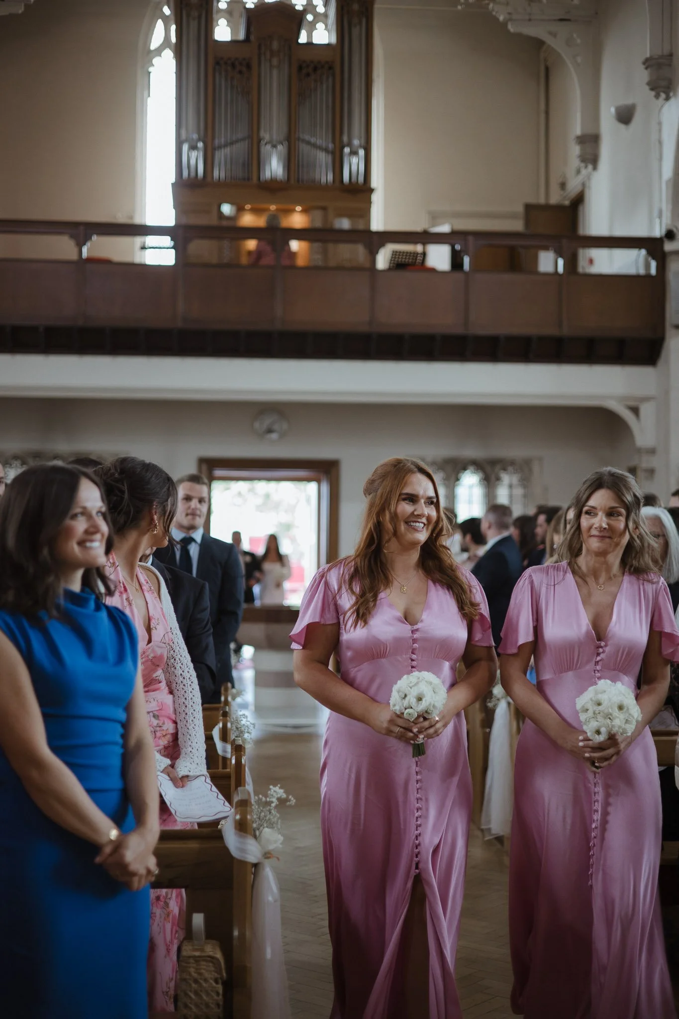Women in pink dresses holding bouquets walking down aisle during a wedding ceremony in a church.