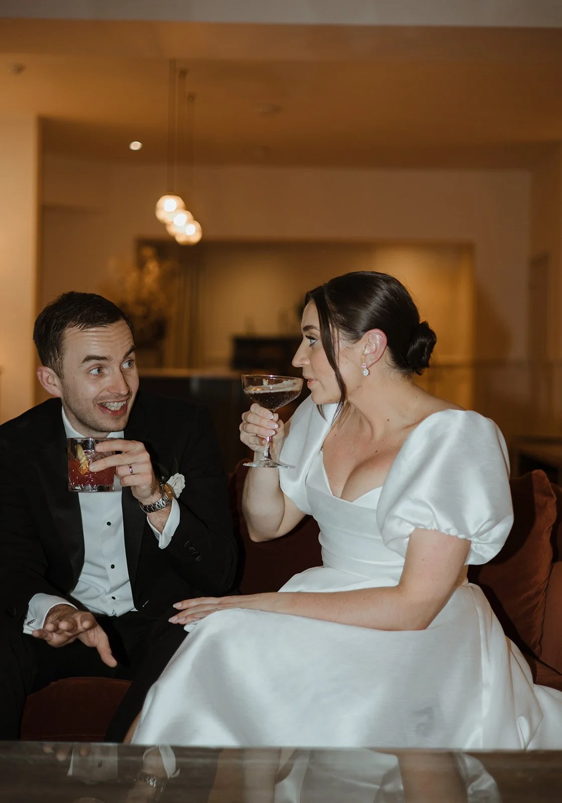 A man in a tuxedo and a woman in a white dress are sitting together, enjoying drinks and engaging in conversation in a warmly lit room.