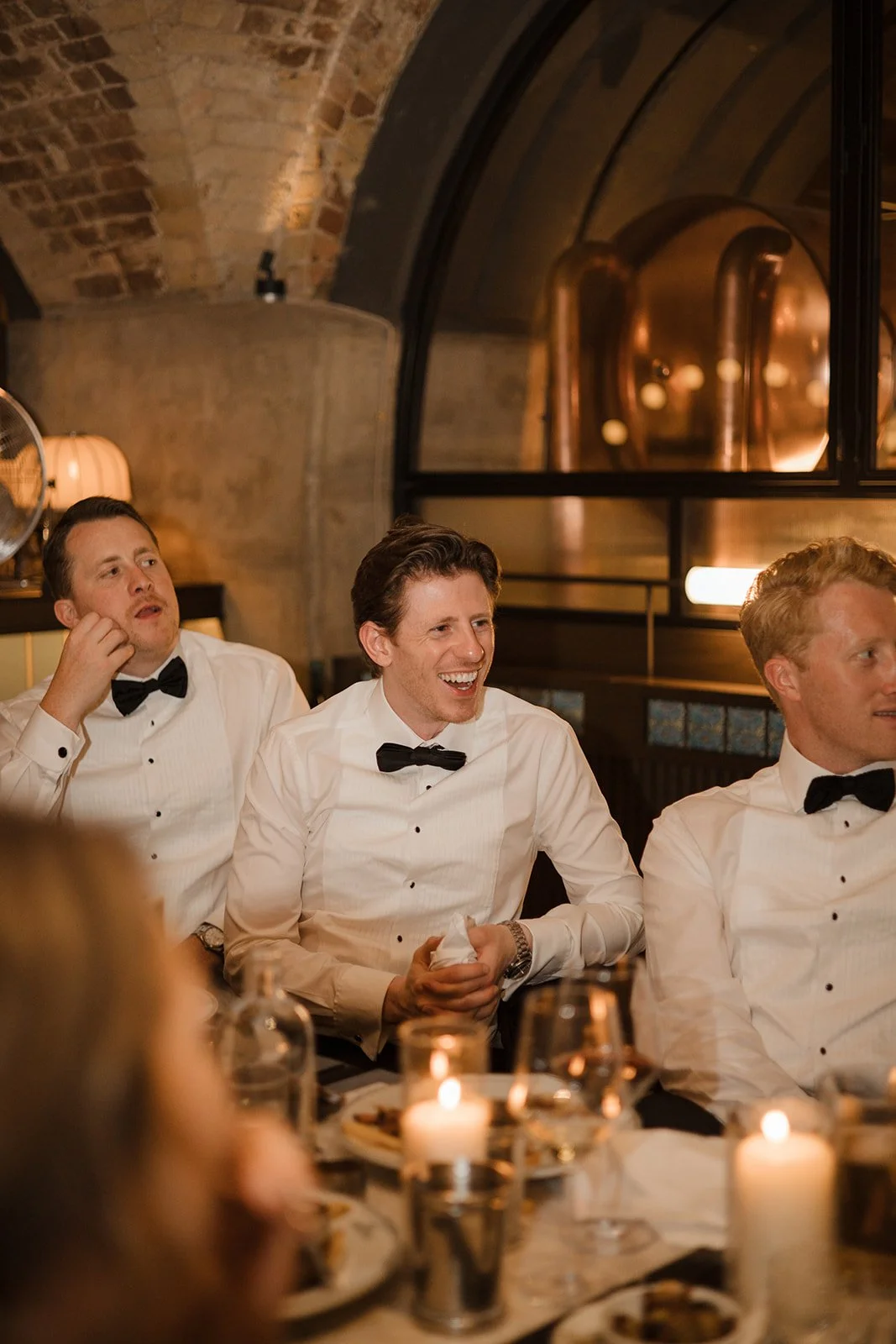 Three men dressed in tuxedos with black bow ties sitting at a formal dinner table with lit candles, enjoying a social event or celebration in a warmly lit, rustic venue with brick arches and large copper brewing tanks in the background.