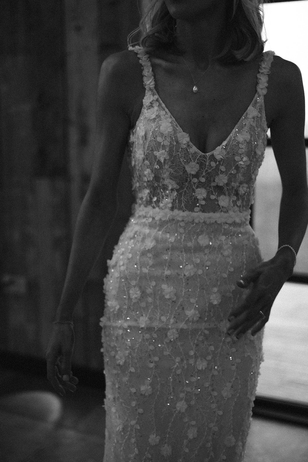 A woman in a lace wedding dress with floral details, wearing a necklace with a pendant, and a bracelet, standing indoors.