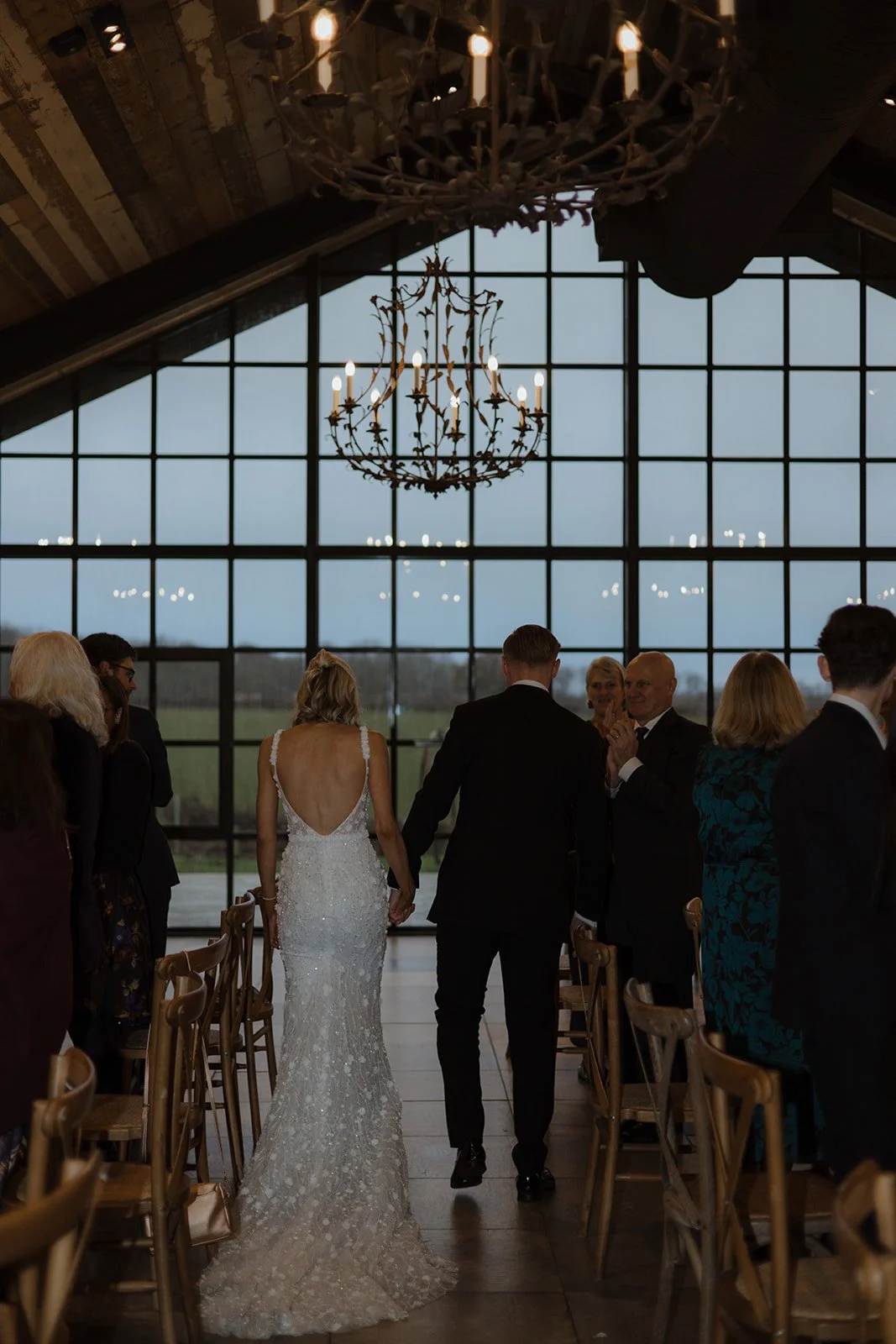A bride and groom walking hand in hand down the aisle during their wedding ceremony, surrounded by seated guests in a spacious, rustic venue with large windows and chandeliers.