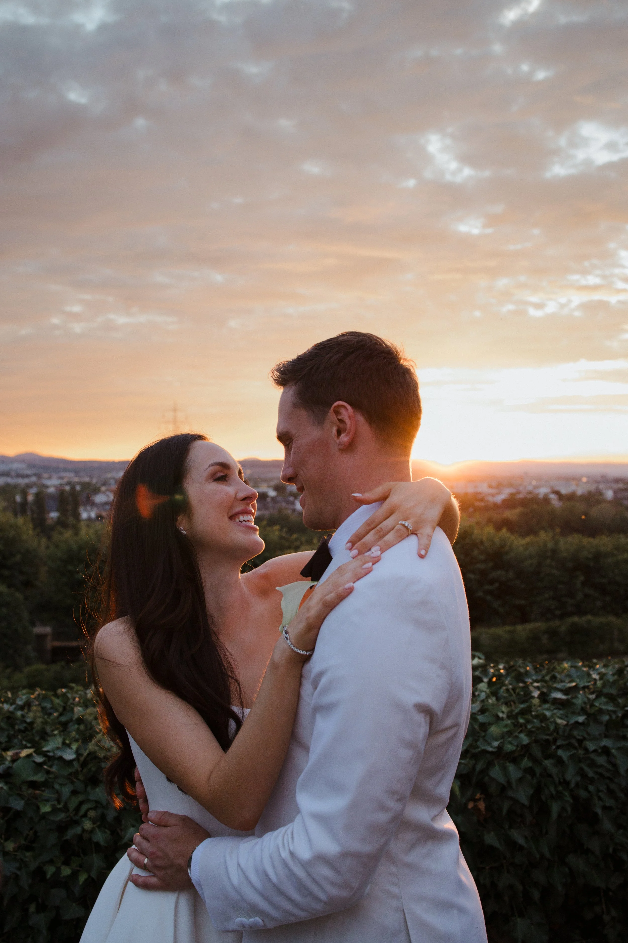 A couple dancing outdoors at sunset, with the woman smiling and the man holding her, against a scenic cityscape and cloudy sky.