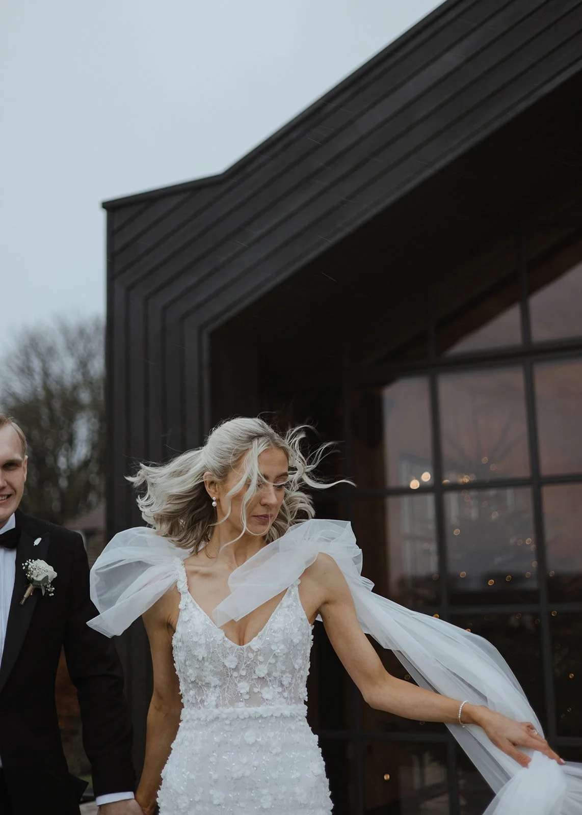 Woman in wedding dress dancing outdoors on a cloudy day with man in tuxedo, black building with large windows in background.