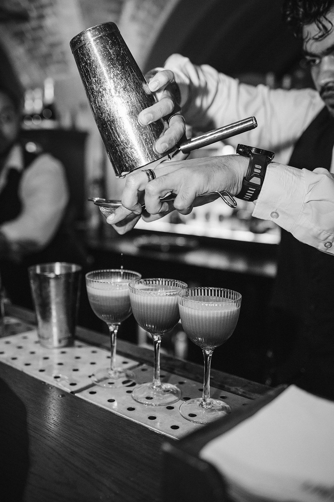 A bartender pours a cocktail through a strainer into three glasses with layered drinks, on a bar counter.