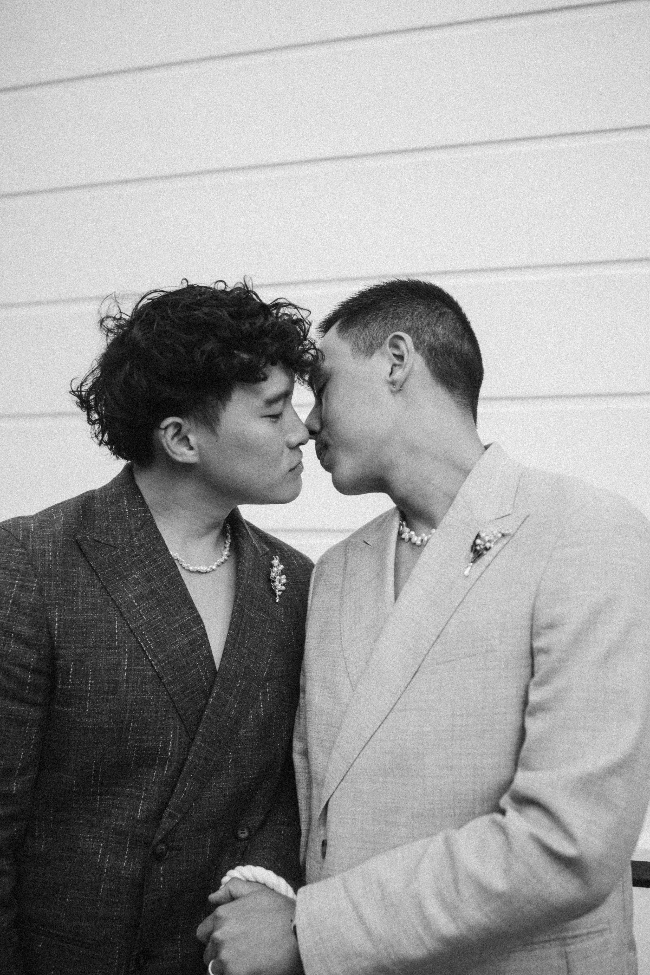 Two individuals dressed in suits are facing each other, holding hands, and with their foreheads touching in a moment of intimacy in front of a light-colored wall with horizontal paneling.