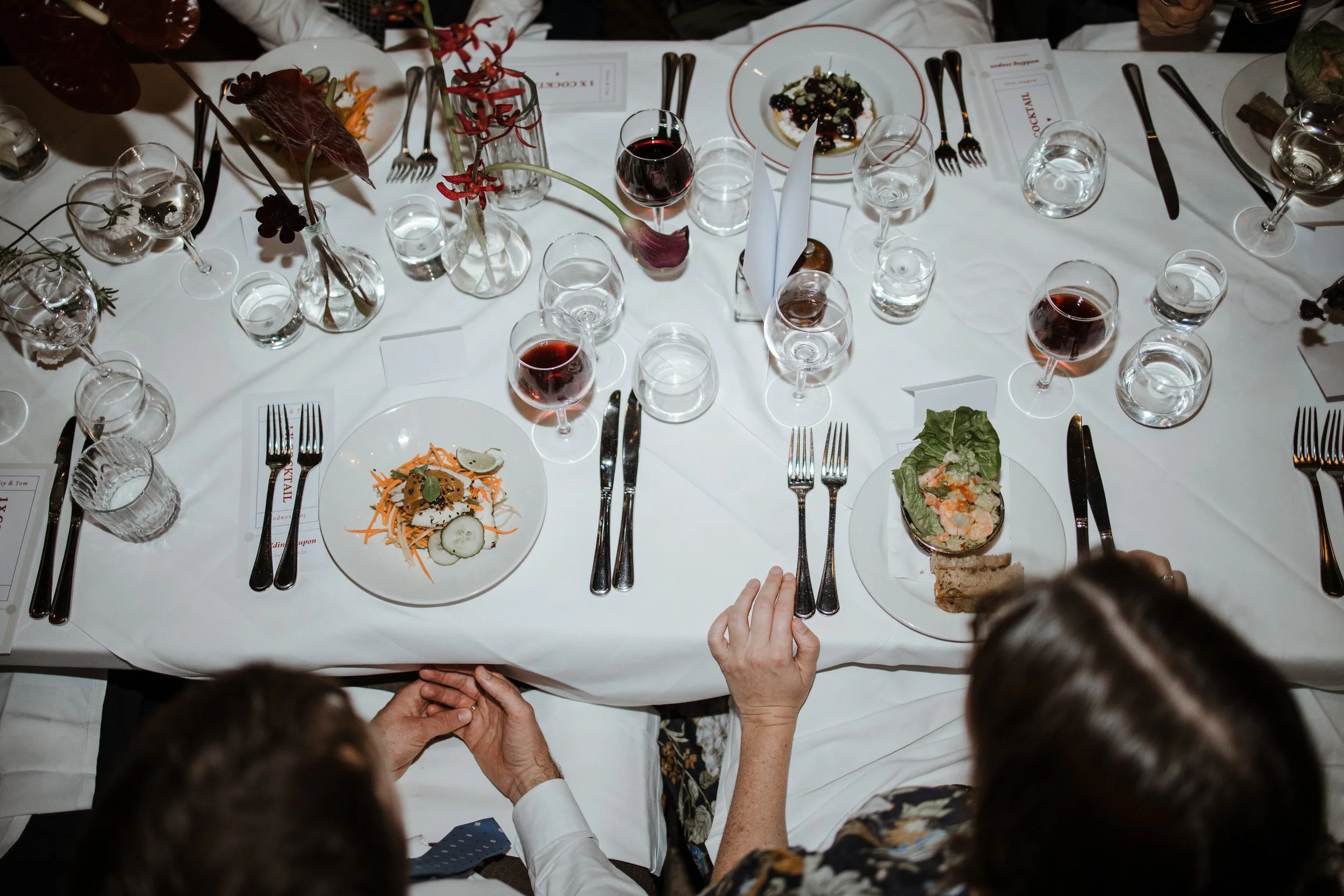 A dinner table set for a formal meal with multiple wine glasses, plates with salads, and silverware. There are also decorative vases with flowers, and some menus and napkins placed on the table.