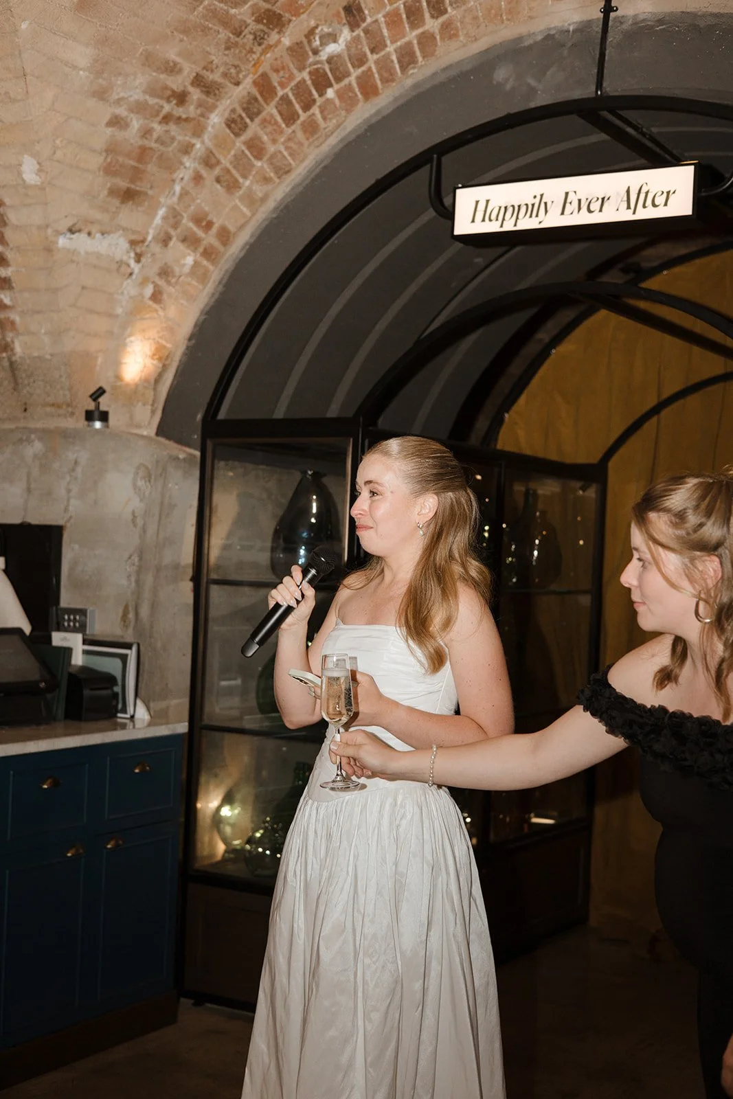 A bride in a white dress giving a speech at her wedding reception, holding a microphone and a glass of champagne. Another woman is handing her a phone. A sign above reads, 'Happily Ever After.'