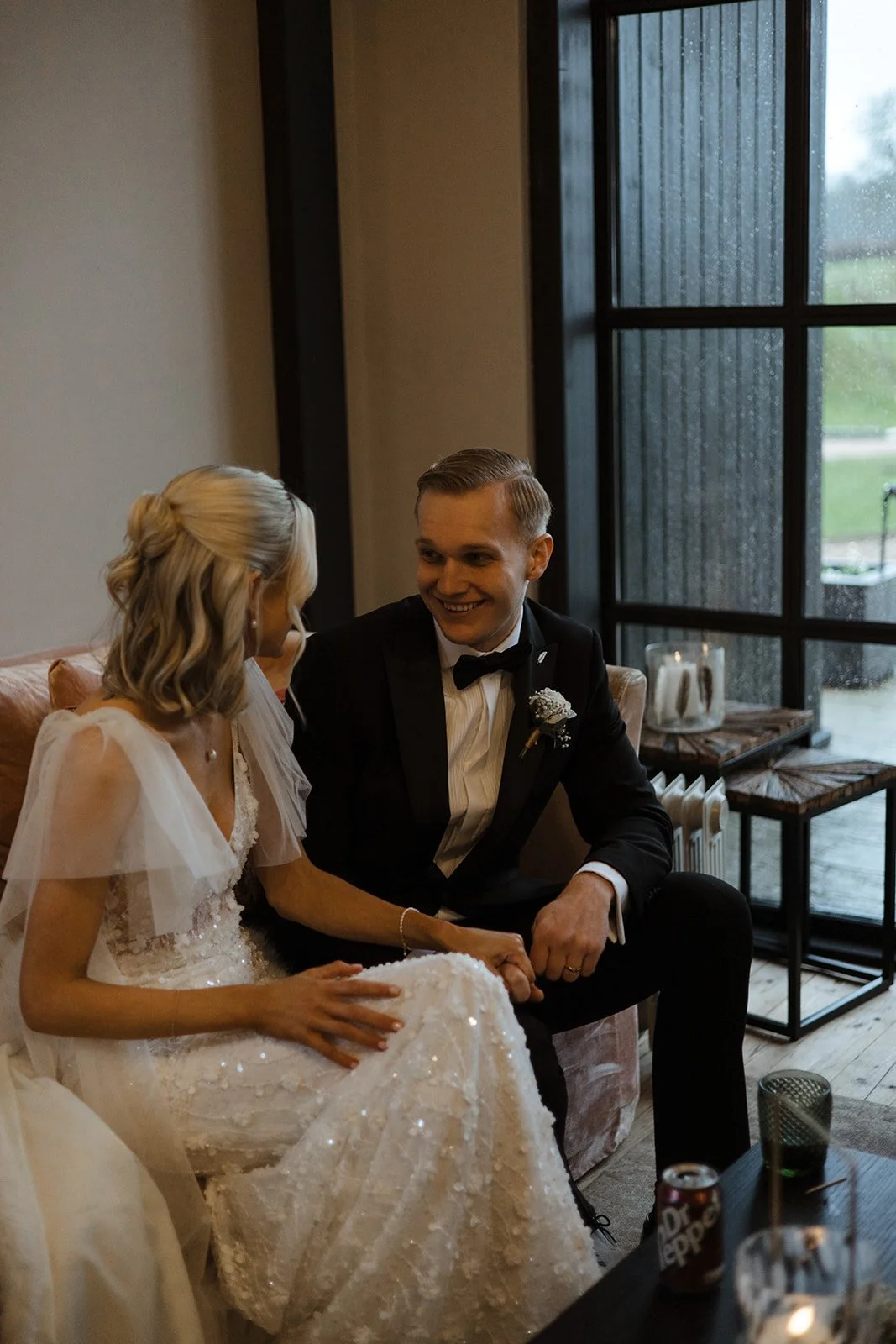 A bride and groom sitting on a sofa, holding hands and smiling at each other, in a room with large windows and wooden floors.