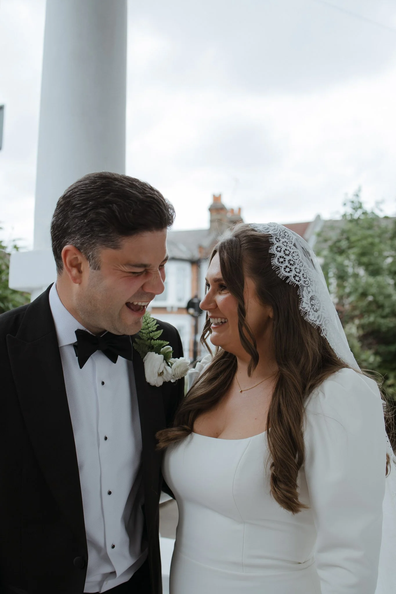 A bride and groom are smiling and laughing together indoors near a window, with the groom wearing a black tuxedo and the bride in a white dress and lace veil.