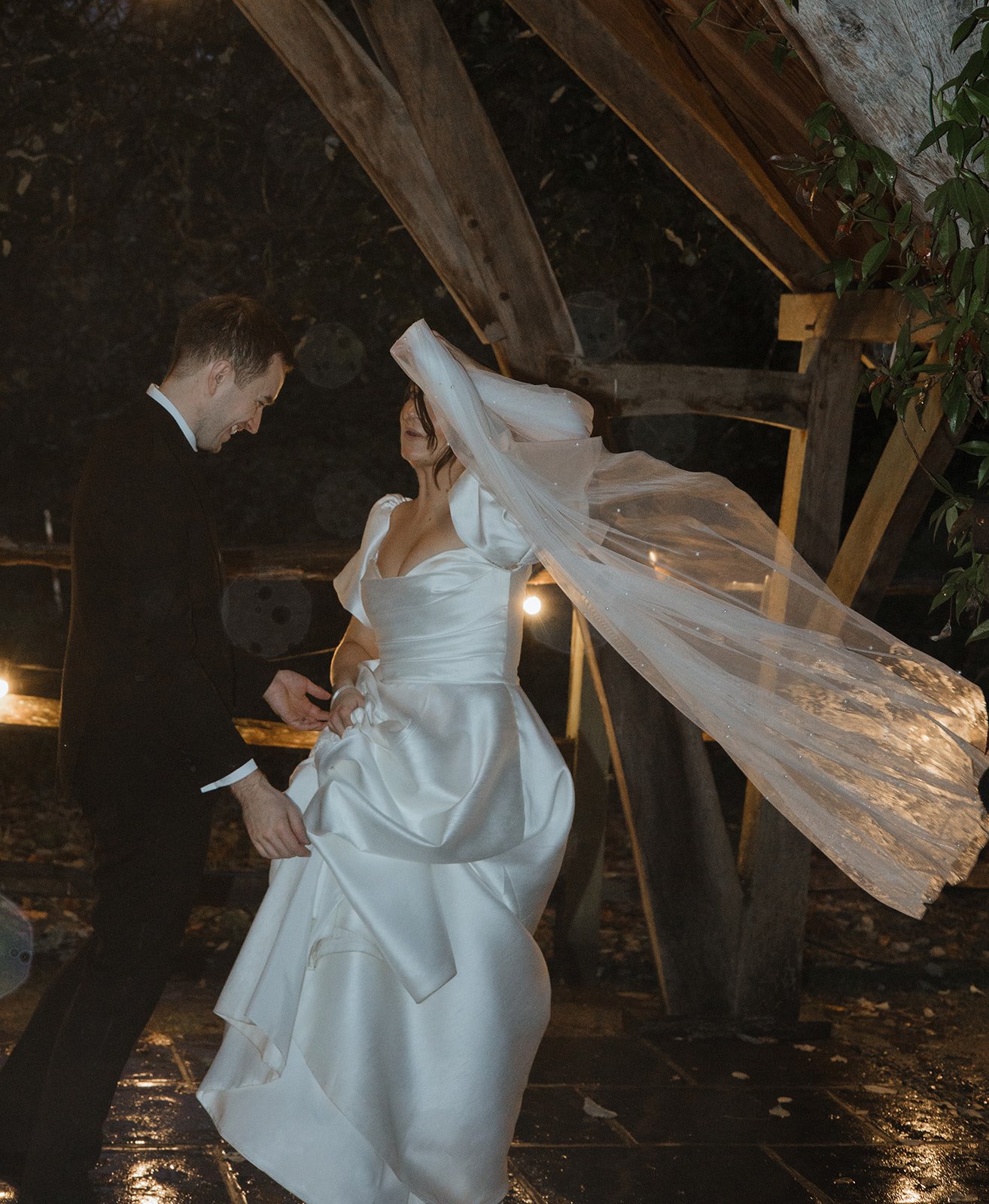 A bride and groom dancing during their wedding reception inside a rustic barn with wooden beams and dim lighting.