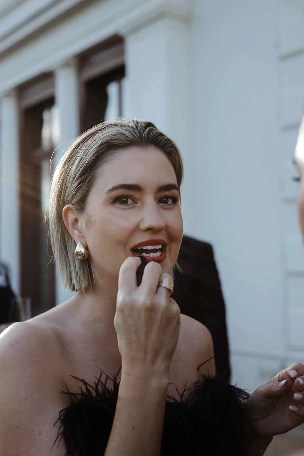 A woman with short blonde hair and gold earrings applying red lipstick outside near a white building.