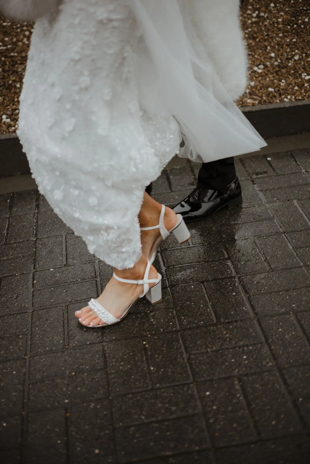 Close-up of a bride and groom's feet, with the bride wearing high-heeled white sandals and the groom in shiny black dress shoes, on a wet brick sidewalk.