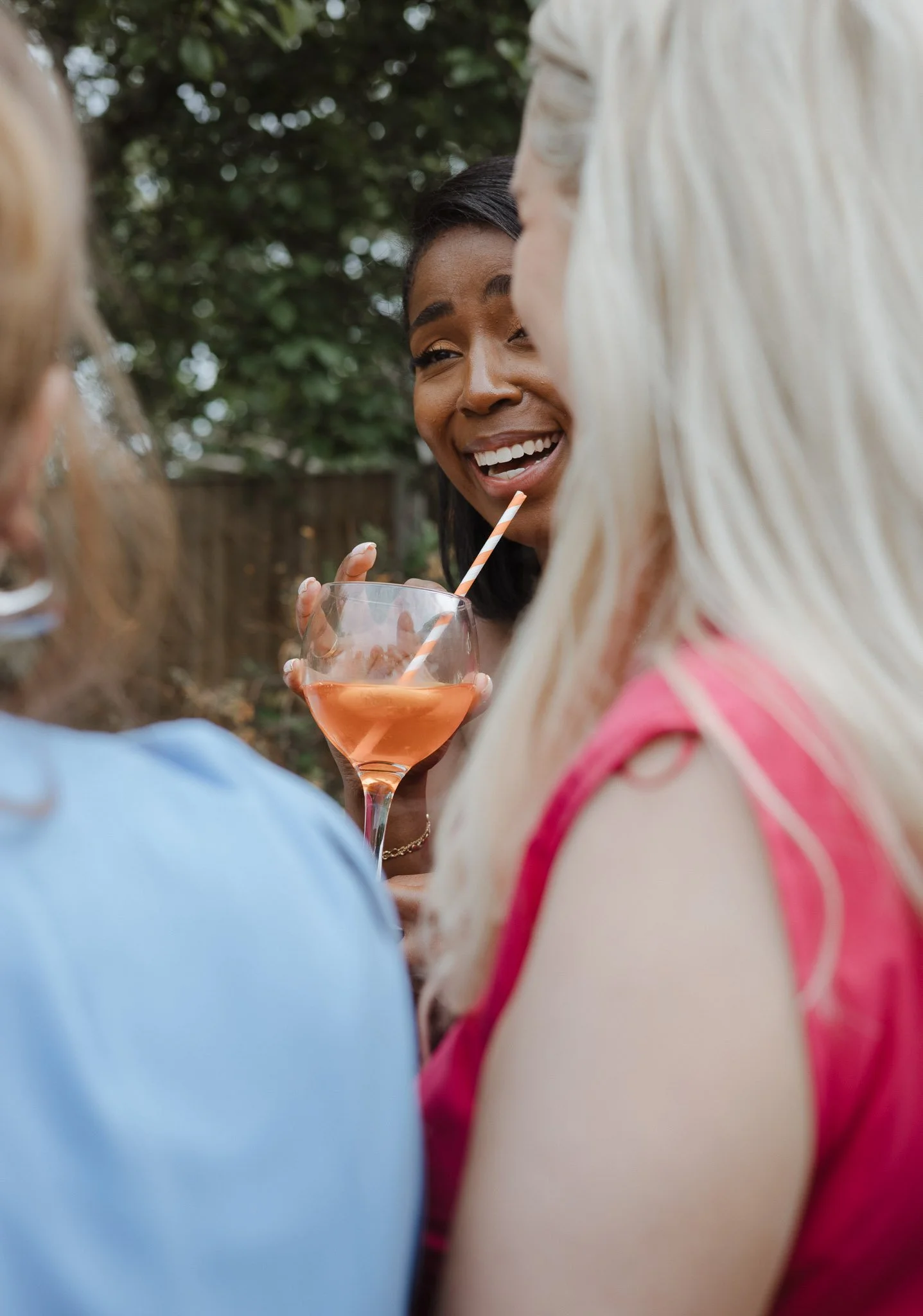 Three women at an outdoor gathering, one holding a pink cocktail with a straw, smiling and engaging in conversation.