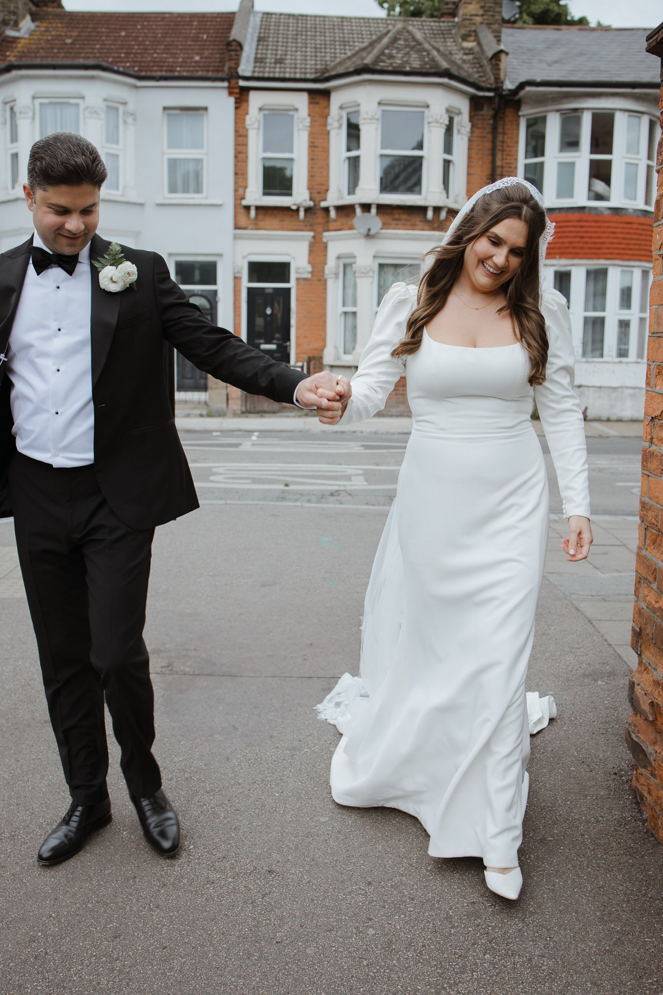 A bride and groom walking hand in hand outside on a city street, smiling, with Victorian-style houses in the background.