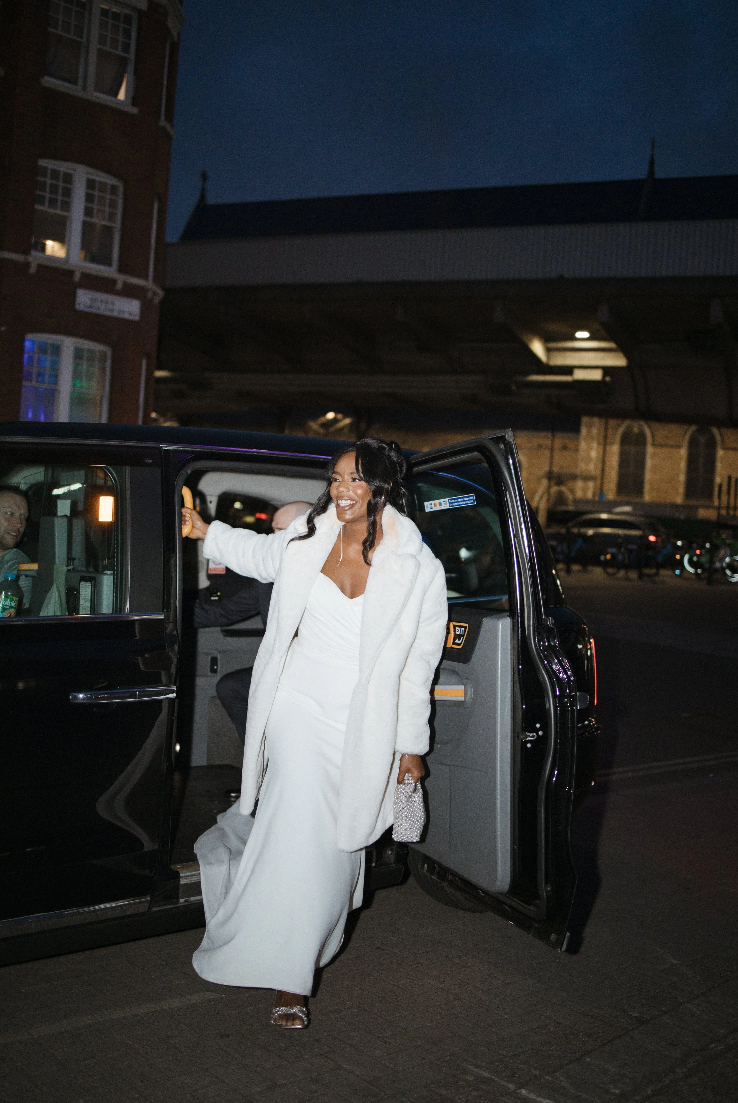 A woman in a white dress and white coat stepping out of a black vehicle at night, holding a small clutch purse, with a broad smile on her face.