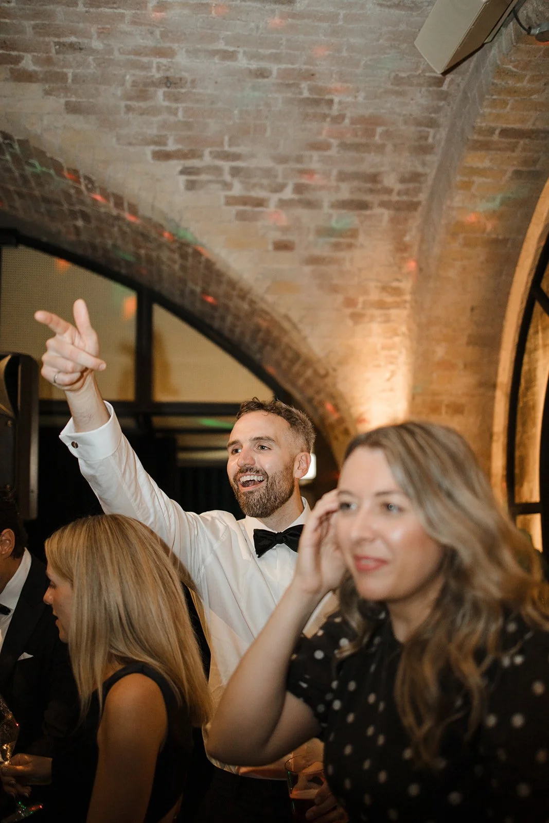 People at a social gathering in a brick-walled venue; a man in a tuxedo with a bow tie is smiling and pointing, and a woman with long wavy hair in a polka dot dress is adjusting her hair.