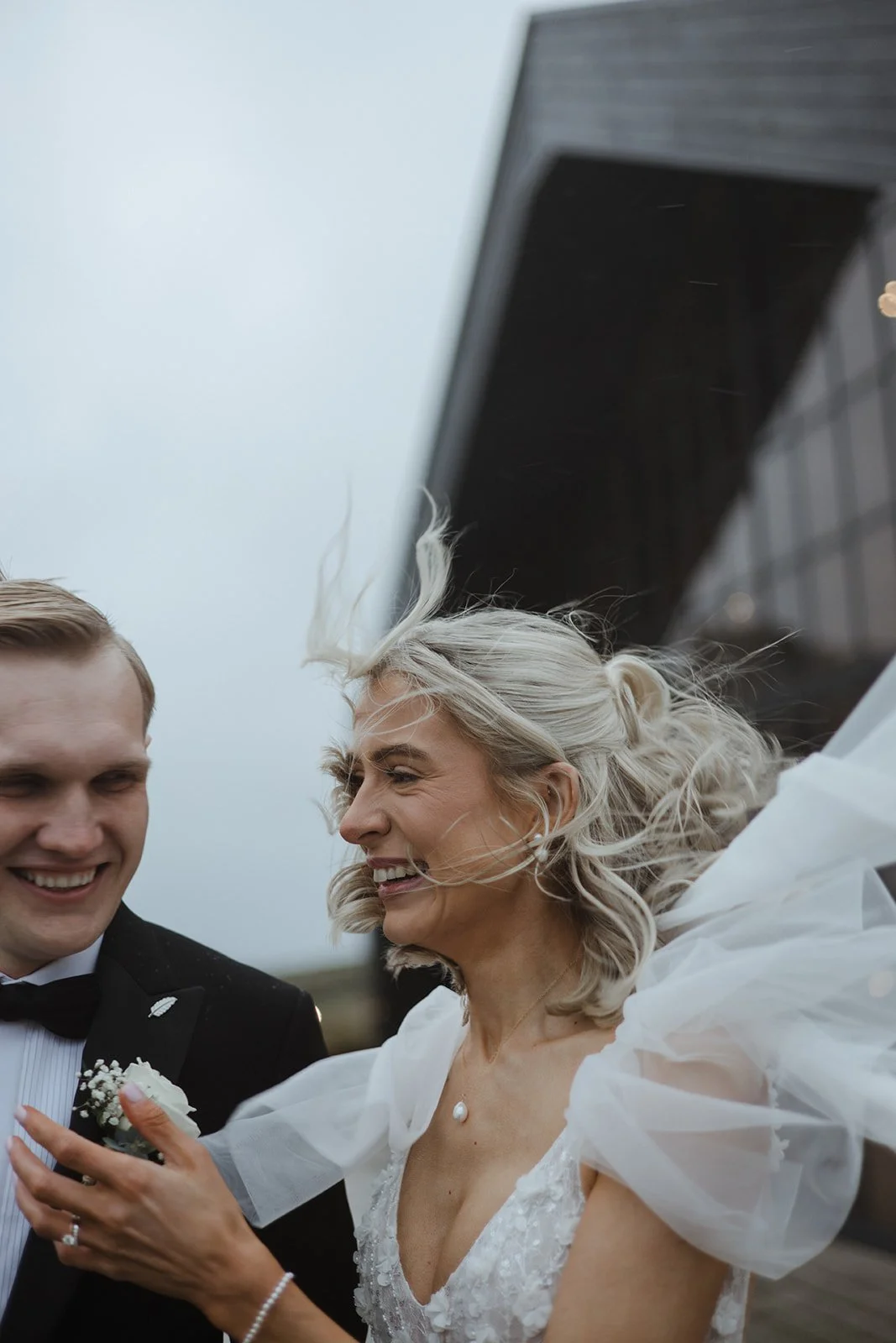 A bride with blonde hair in a white wedding dress and a groom in a black tuxedo with a bowtie smile and laugh outdoors, with wind blowing their hair and a building in the background.
