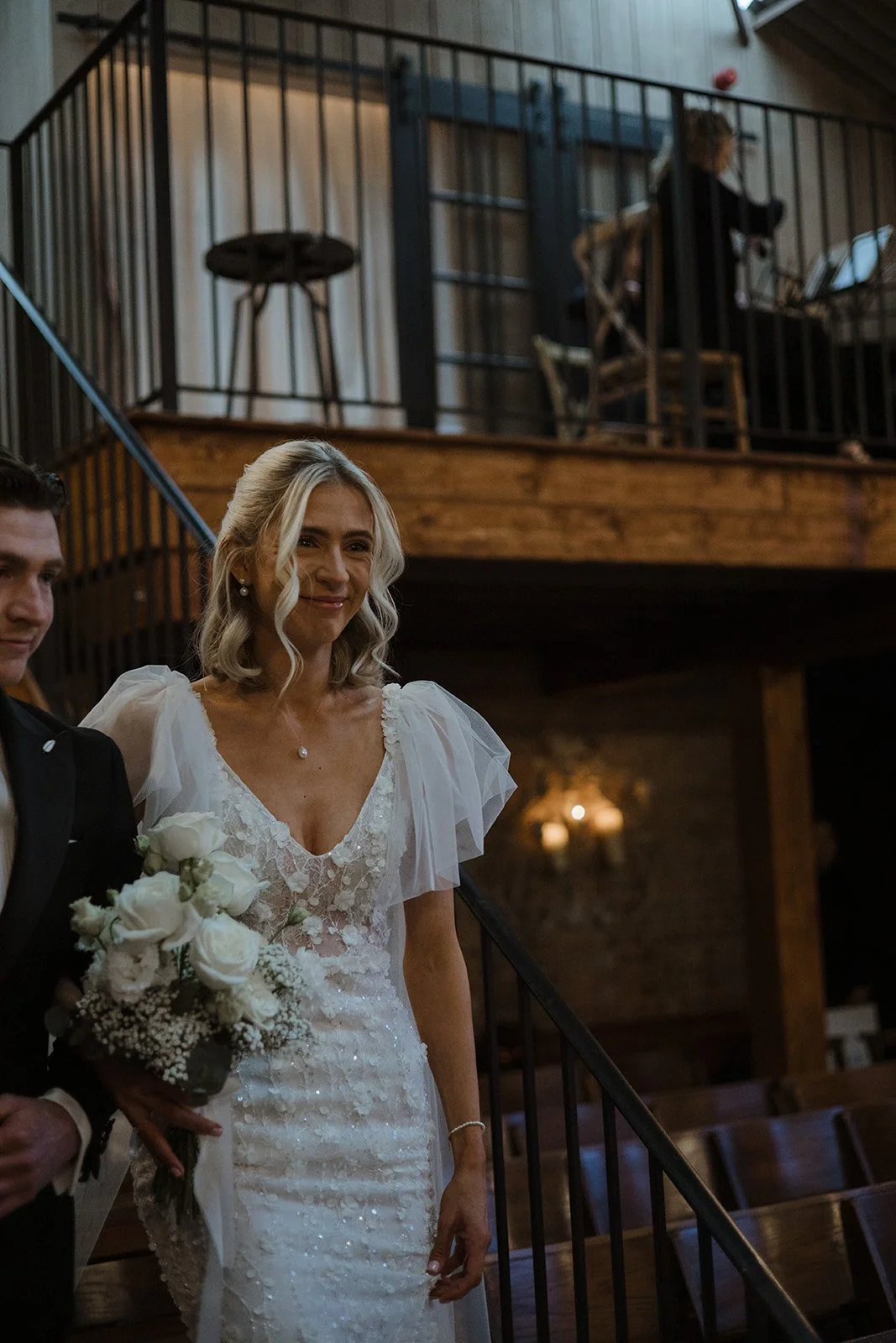 A bride holding a bouquet of white roses, wearing a lace wedding dress with puffed sleeves, standing on a staircase at a wedding ceremony.