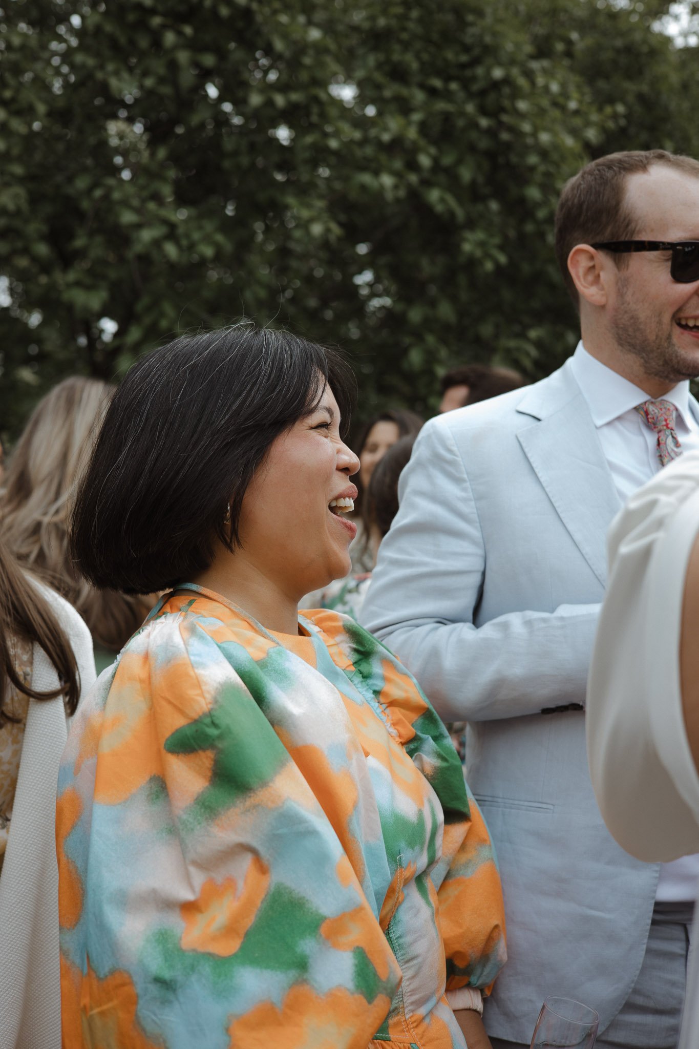 A woman with black hair laughing and smiling at a social outdoor event, wearing a colorful abstract-patterned jacket, with greenery and other people in the background.