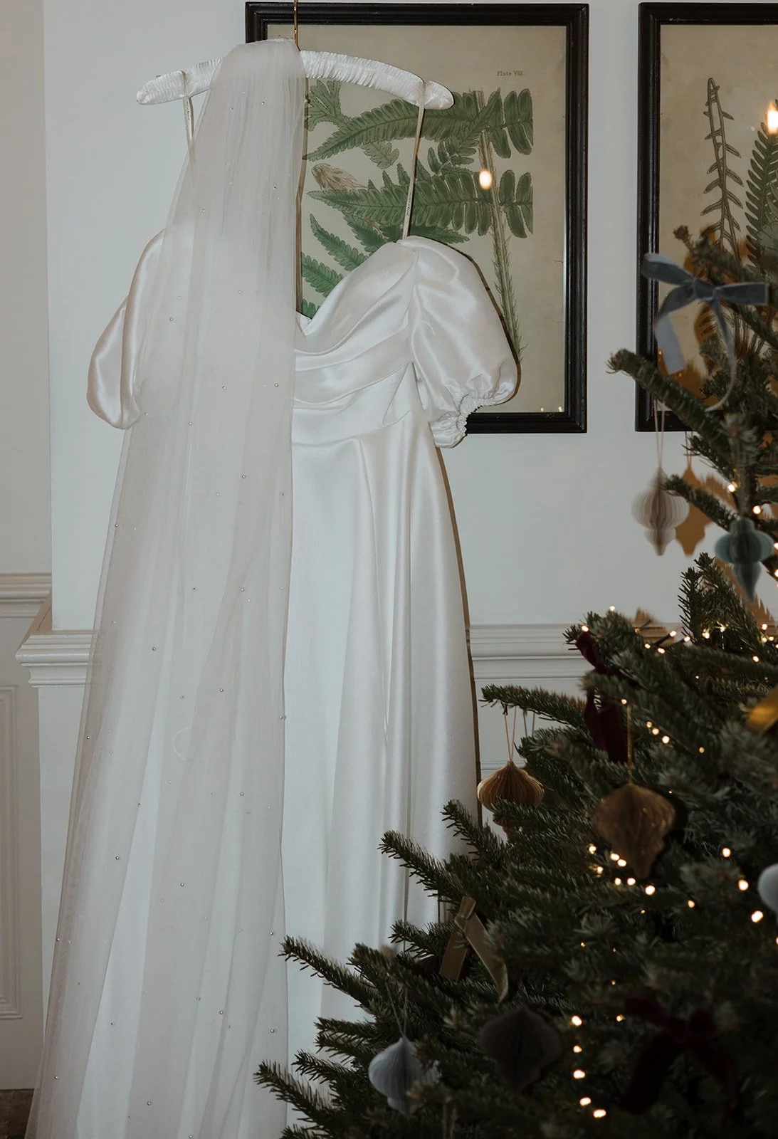 A white wedding dress with puffed sleeves hanging on a black frame behind a Christmas tree decorated with ornaments and lights, with framed botanical prints on the wall in the background.