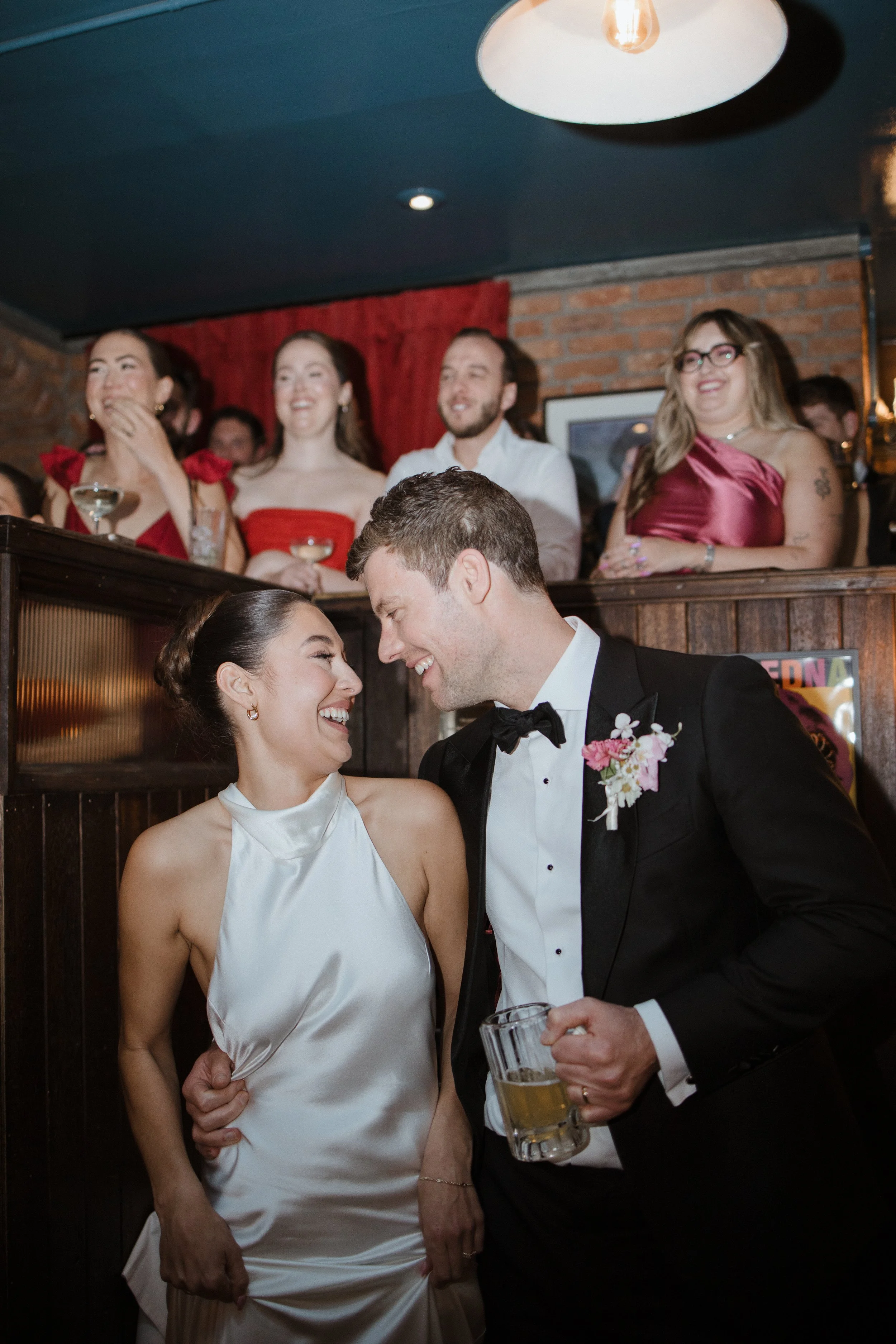A bride and groom are smiling and sharing an intimate moment at their wedding reception, with the bride wearing a white satin dress and the groom in a tuxedo holding a beer mug. Behind them, guests are standing and smiling, dressed in formal attire, in a cozy venue with brick walls and hanging lights.
