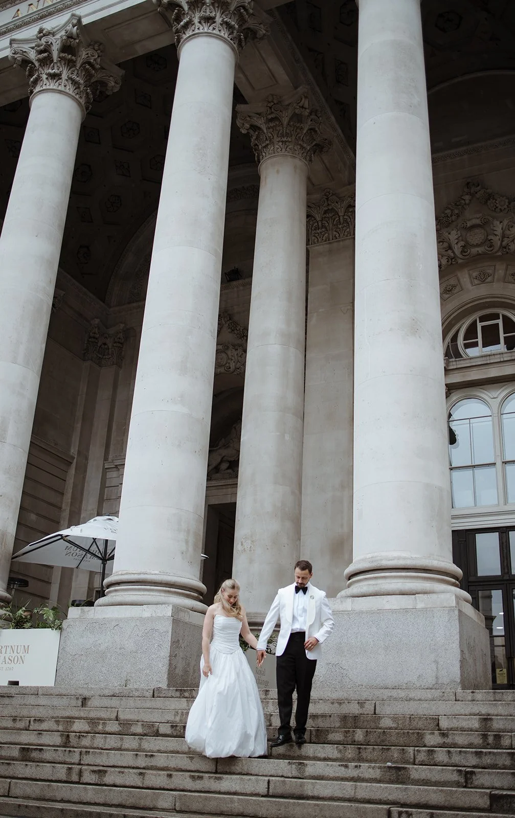 A couple dressed in wedding attire walking down the steps of a large, classical building with tall, ornate columns and arched windows.