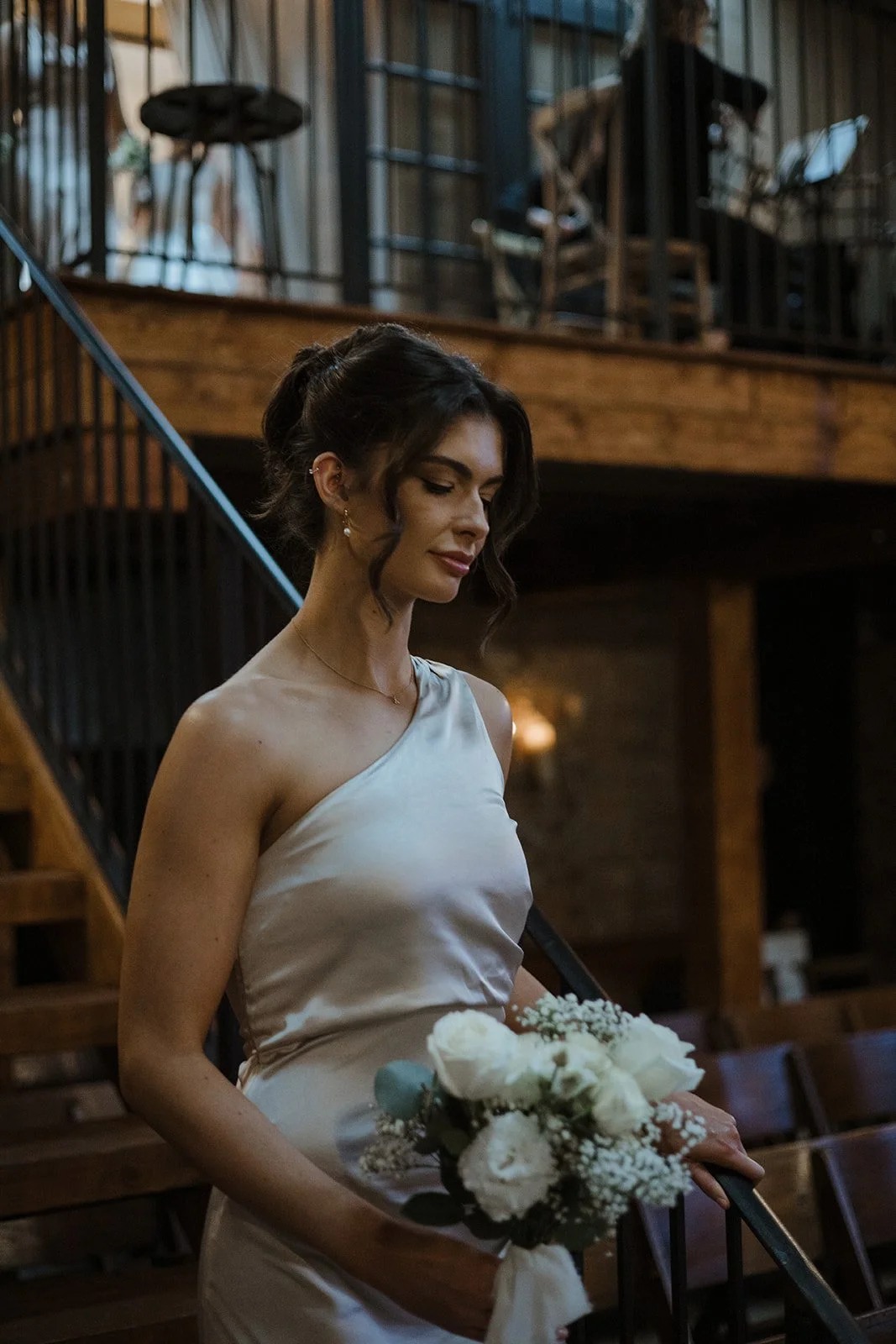 A woman in an elegant, satin, sleeveless dress holding a bouquet of white flowers, standing on a wooden staircase in a warmly lit, rustic indoor setting.