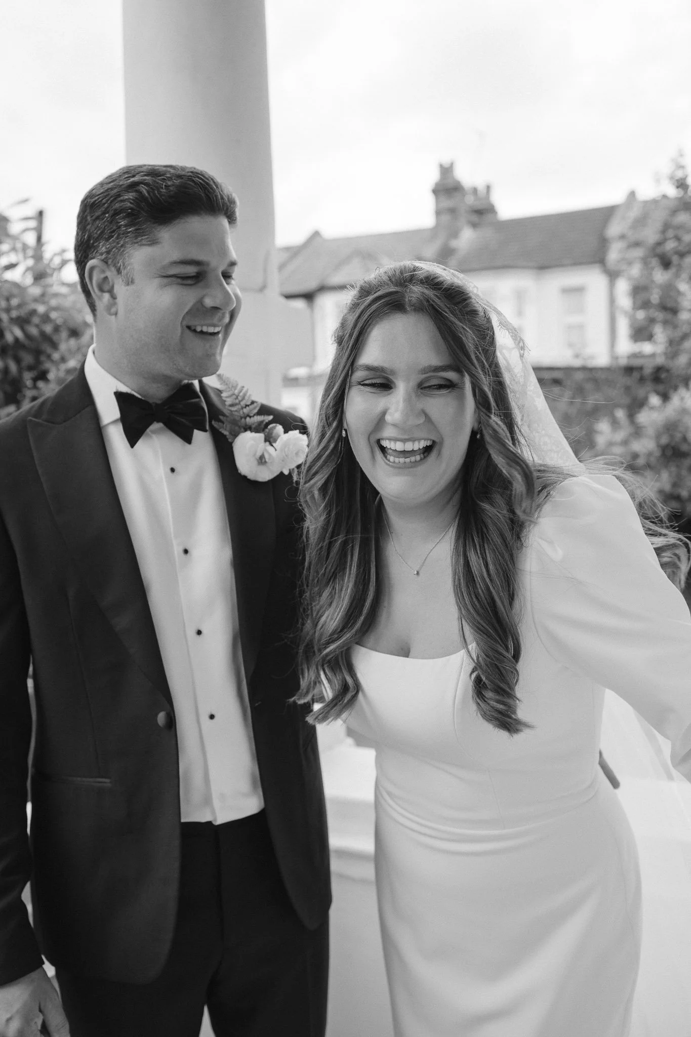 Black and white photo of a man in a tuxedo with a bow tie and a woman in a white dress smiling, standing outdoors.