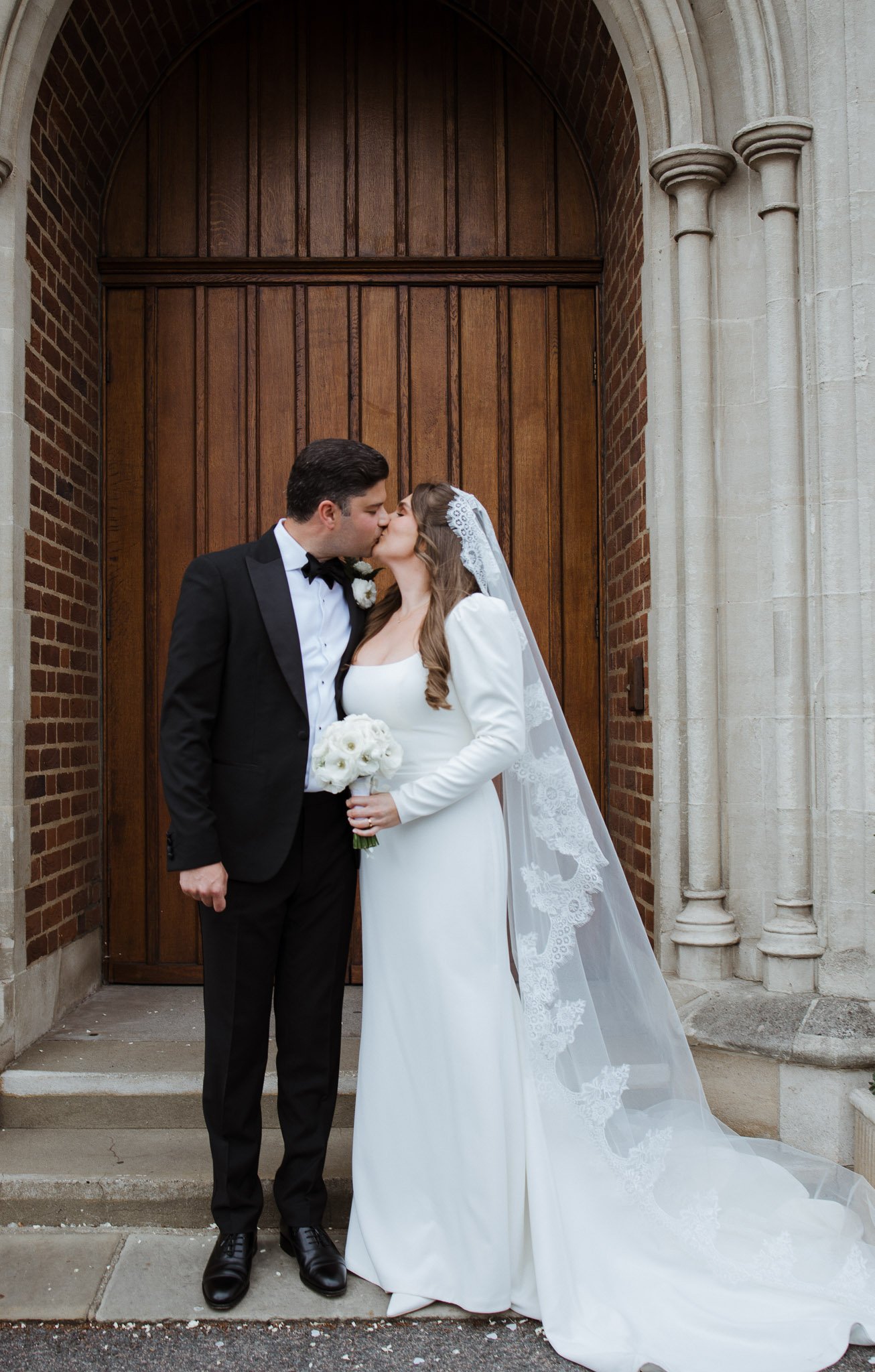 A bride and groom share a kiss outside a church, standing in front of a large wooden door. The bride wears a white dress with long sleeves and a lace veil, holding a bouquet of white roses. The groom wears a black tuxedo with a bow tie.