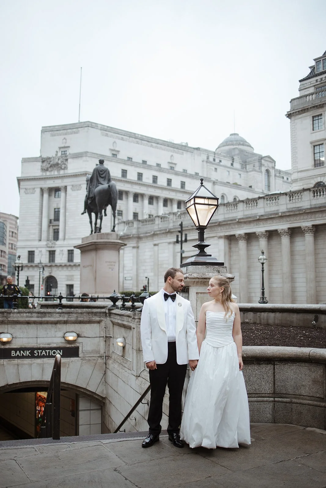 A bride and groom standing together on a city street near a bank station entrance with an American-style governmental building and statue in the background, during daytime with overcast skies.