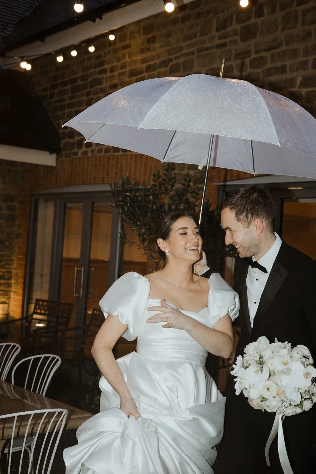 A bride and groom smiling under an umbrella during their wedding reception, with the bride wearing a white dress and holding a bouquet of white flowers, and the groom in a black tuxedo, in a rustic outdoor setting with brick walls and string lights.