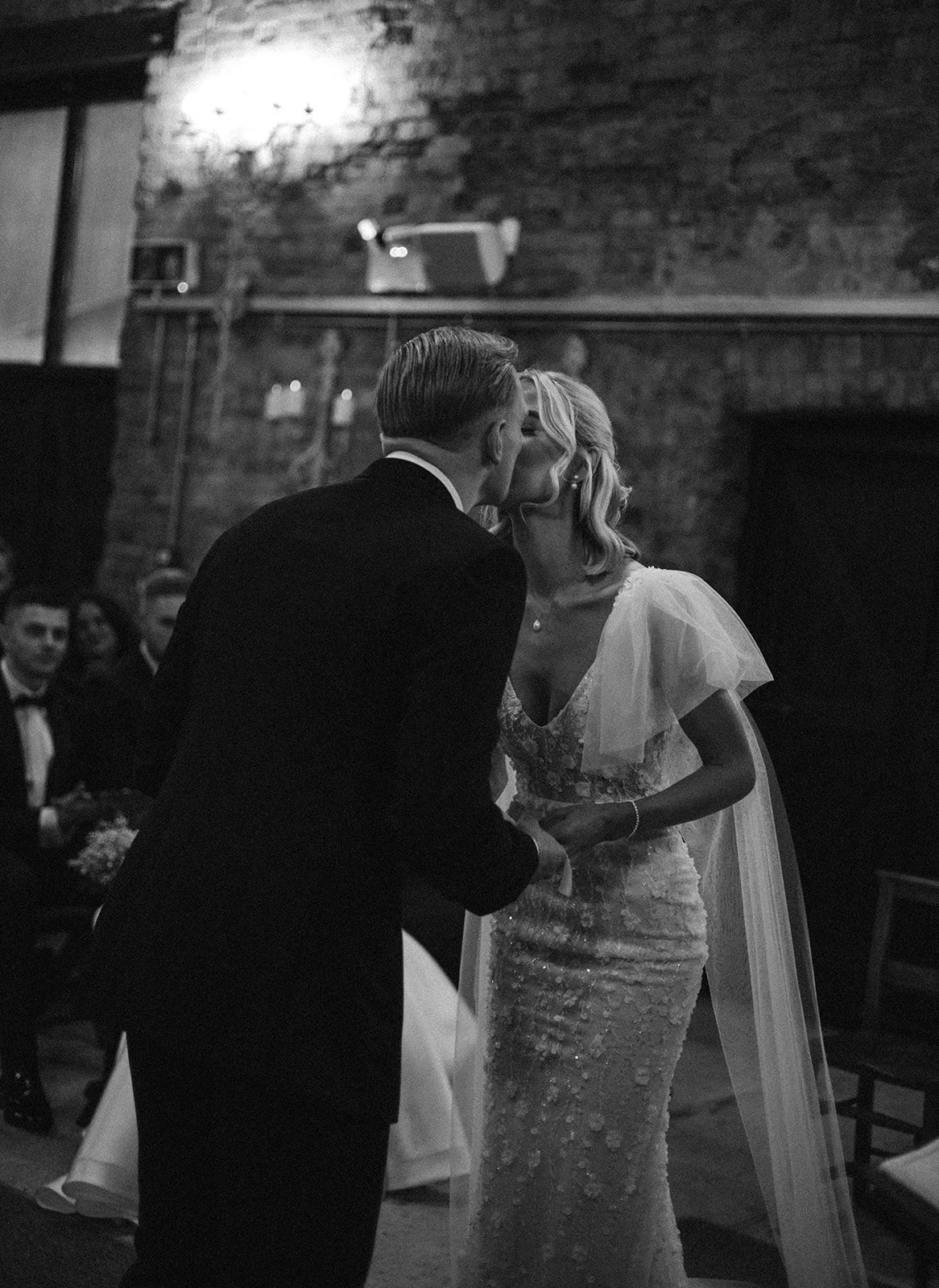 A bride and groom sharing a kiss during their wedding ceremony in a rustic indoor venue with exposed brick walls and guests watching.