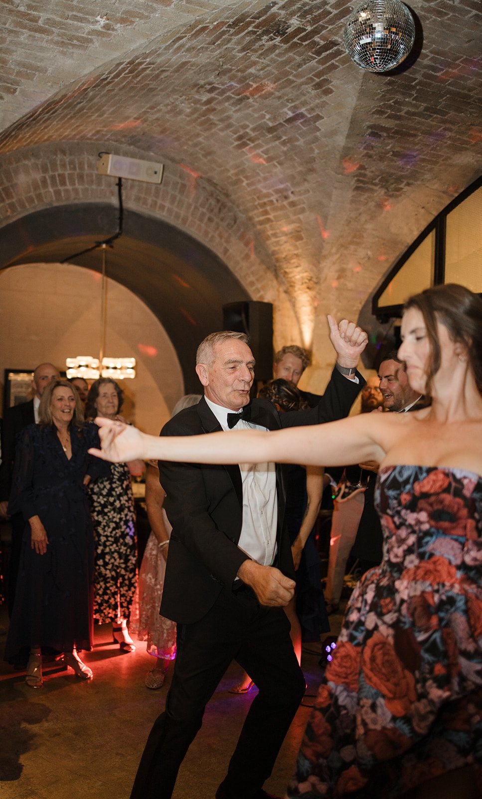 Older man in a tuxedo dancing with a young woman in a strapless dress at a party. Other people are in the background, smiling and enjoying the event in a brick-walled venue with a disco ball overhead.