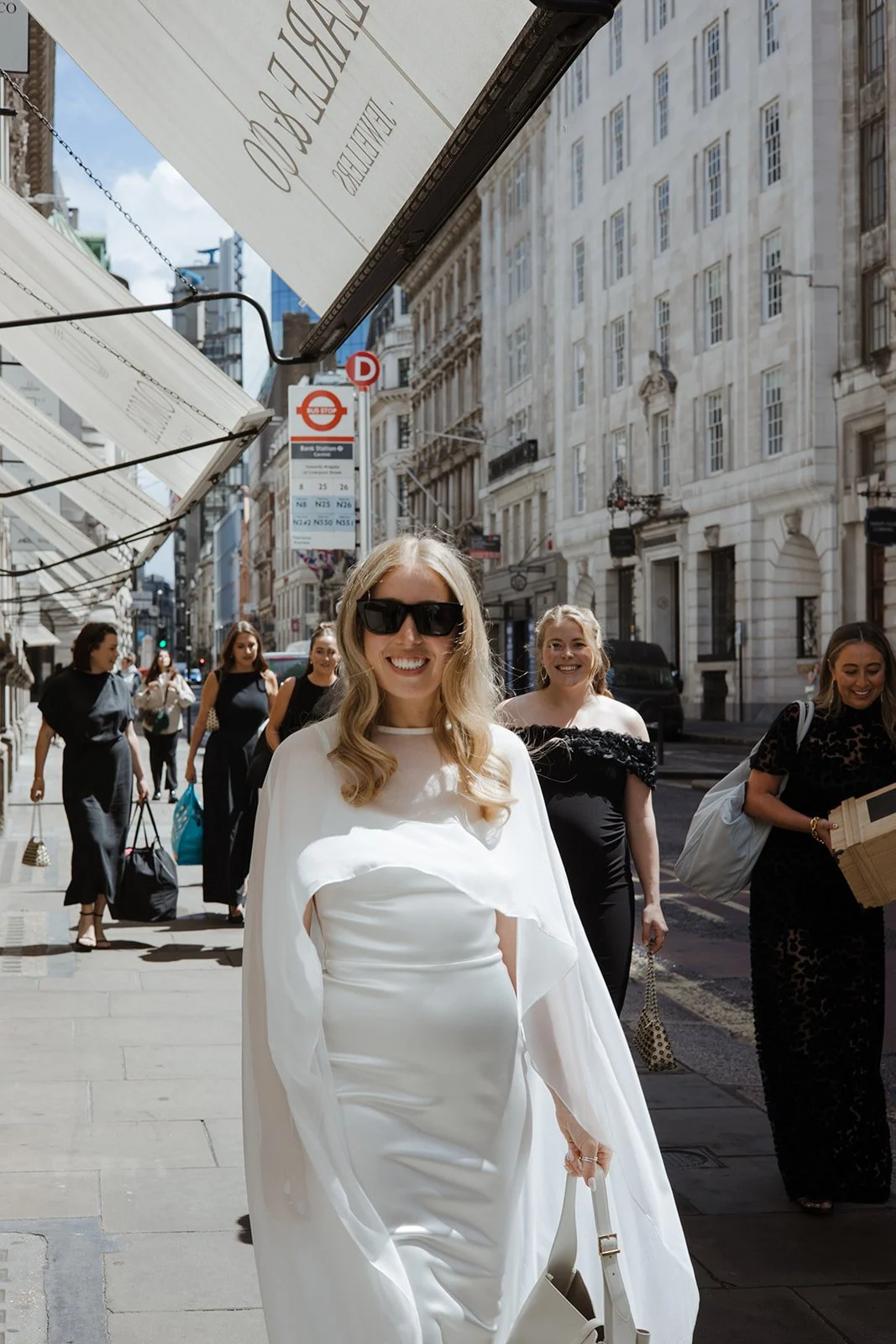 A group of women walking down a city street, with one woman in a white dress and sunglasses smiling at the camera.