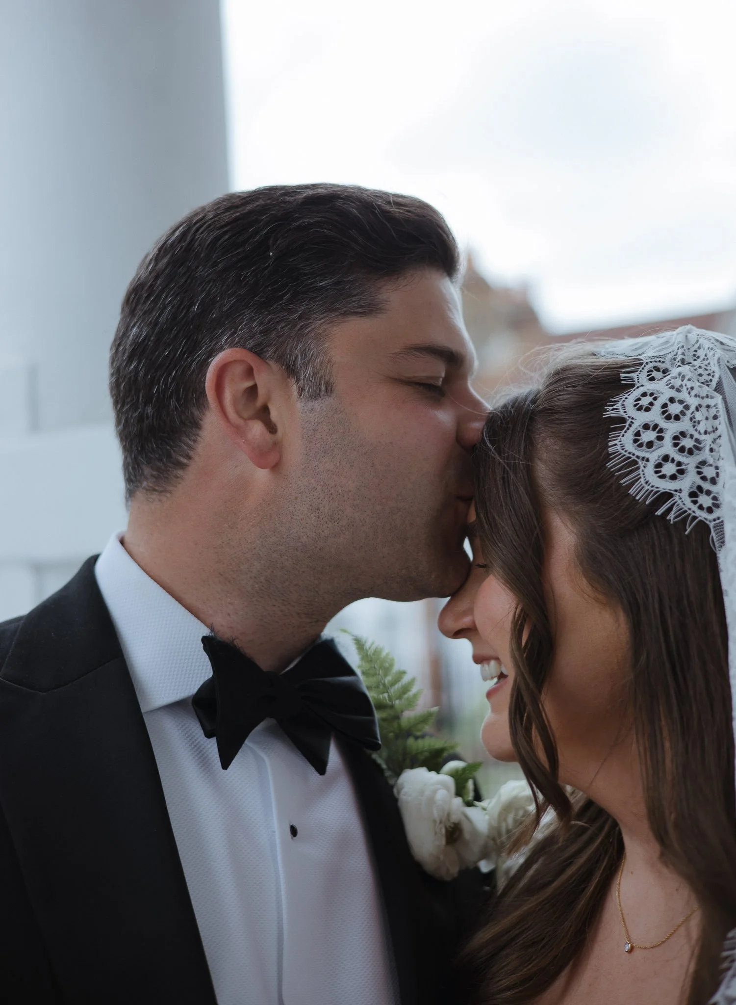 Groom kissing bride on forehead during wedding ceremony, bride wearing lace veil, groom in tuxedo with bowtie, smiling.