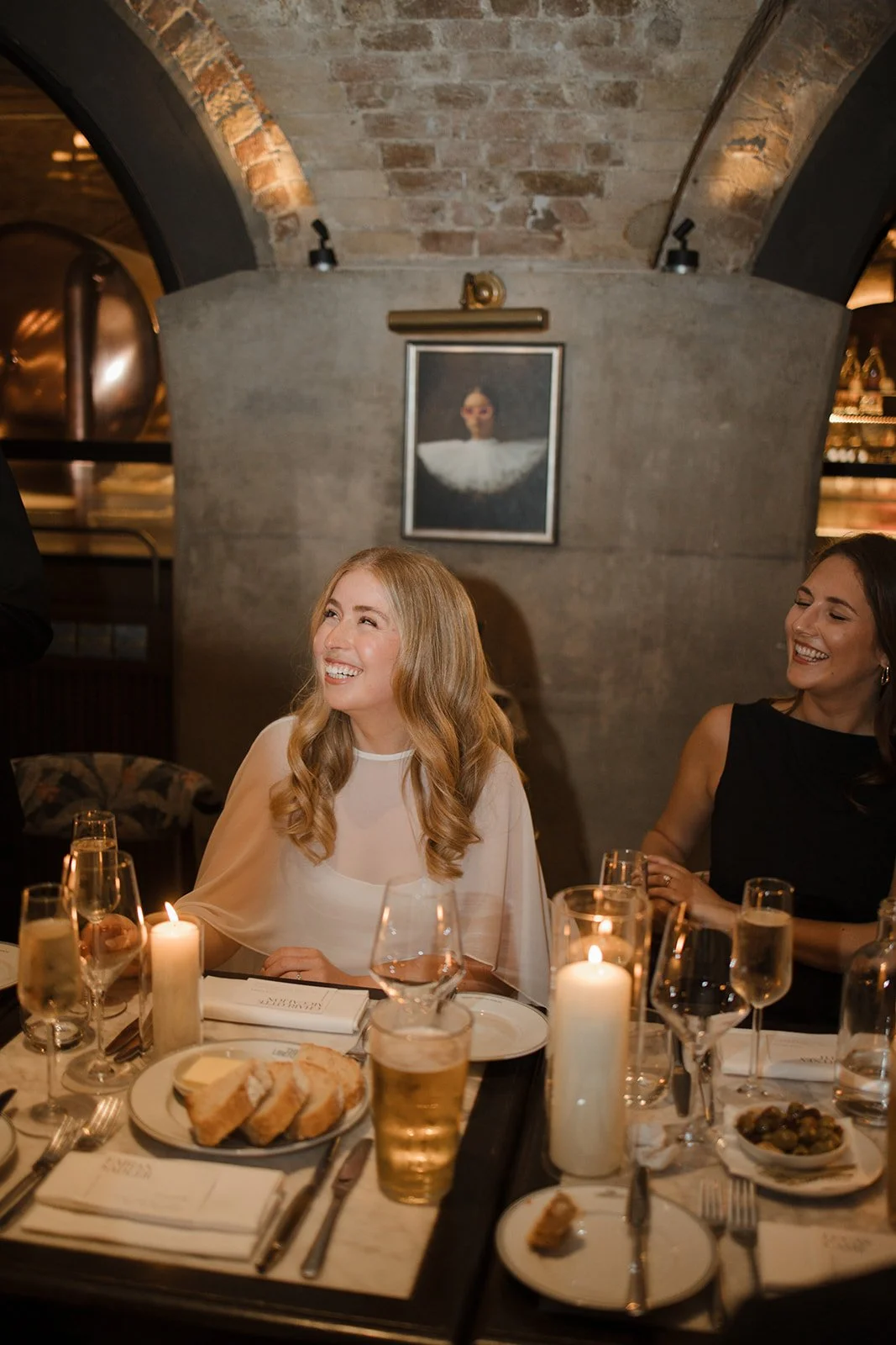Two women are seated at a dinner table, smiling and laughing. The table is set with candles, glasses of wine, plates of food, and utensils. The background features a brick arch ceiling and a framed portrait on a stone wall.