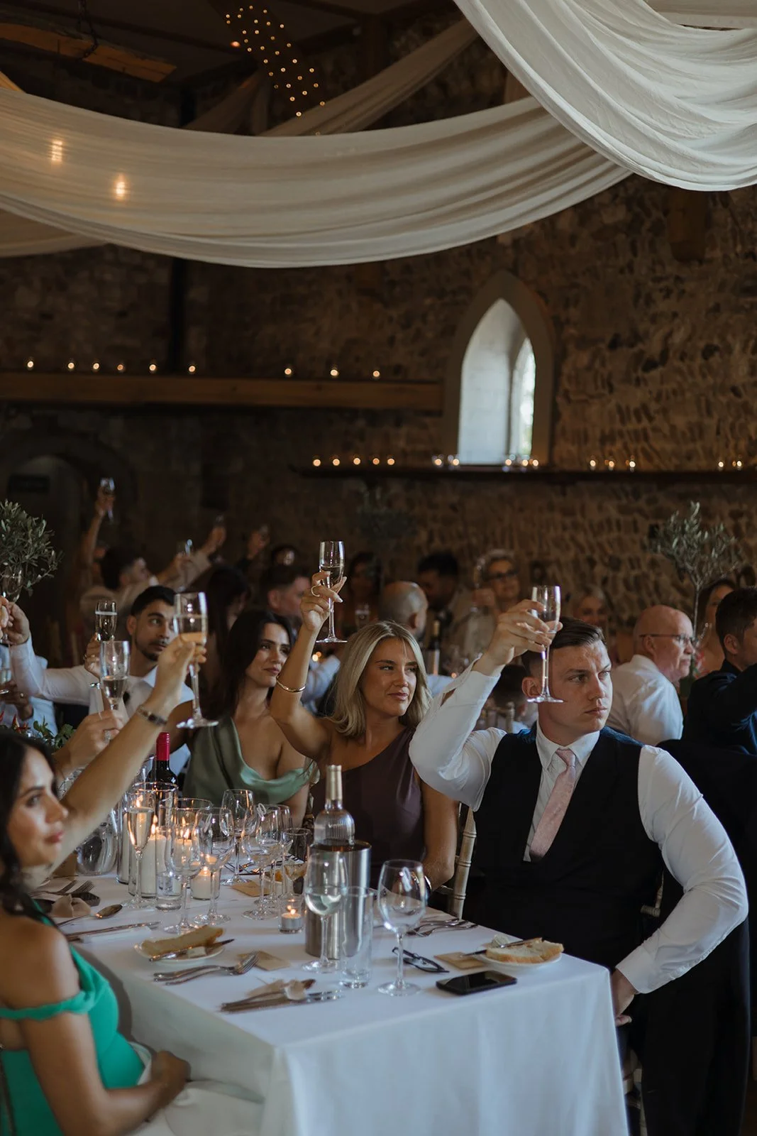People raising glasses in a toast at a wedding reception inside a stone building with draped white fabric on the ceiling.