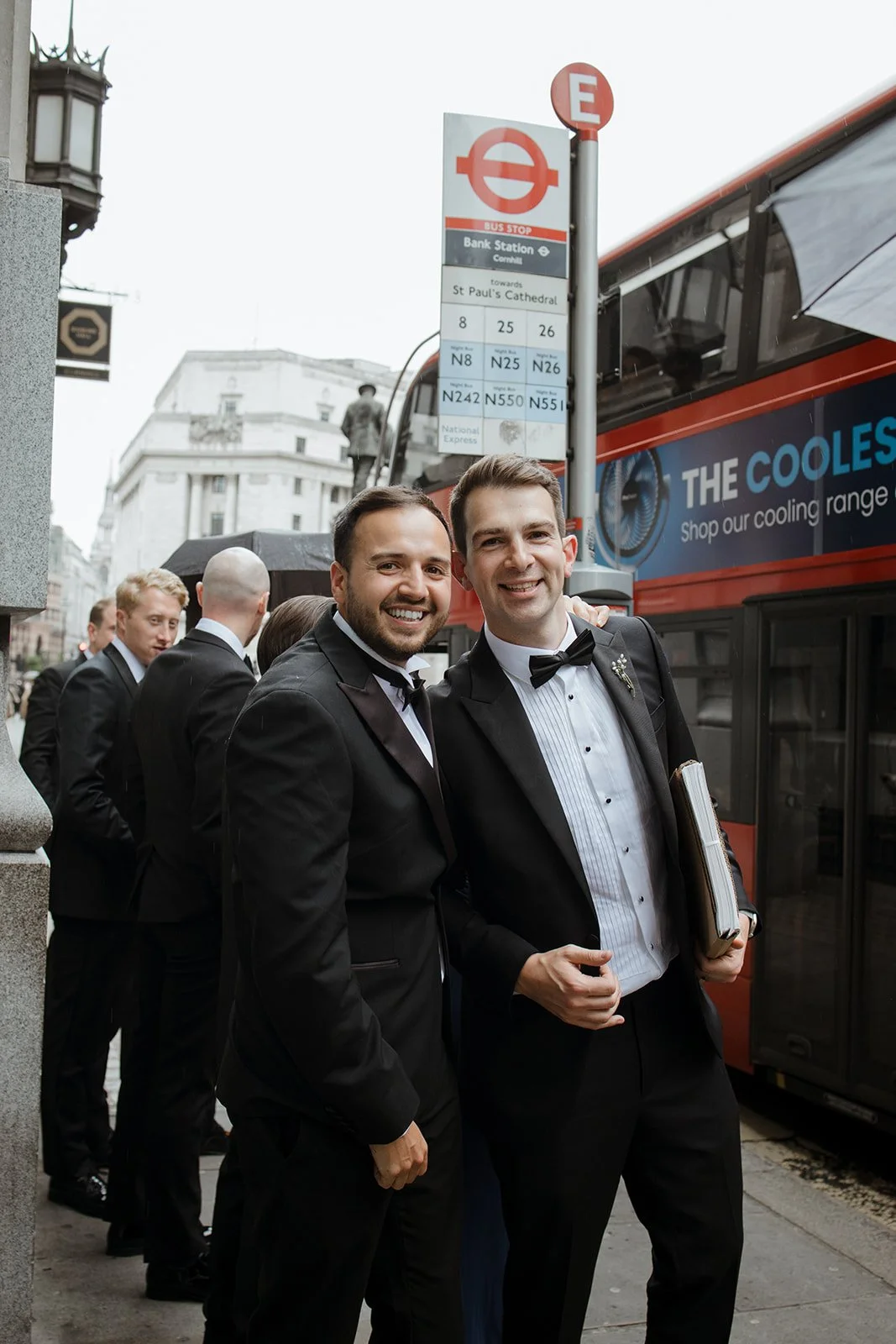 A group of men in tuxedos standing on a city street during daytime, smiling and posing for a photo, with a red double-decker bus and bus stop signs in the background.