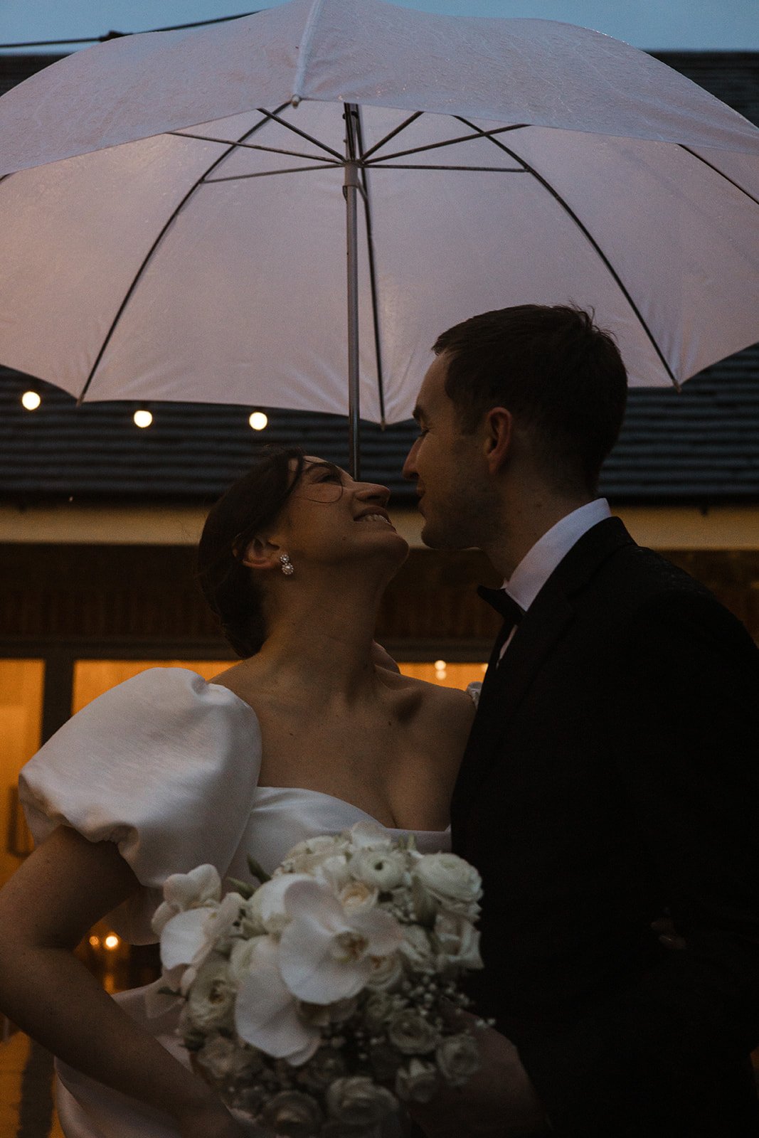 A bride and groom smiling at each other under a large white umbrella during their wedding celebration at dusk, with the bride holding a bouquet of white flowers.
