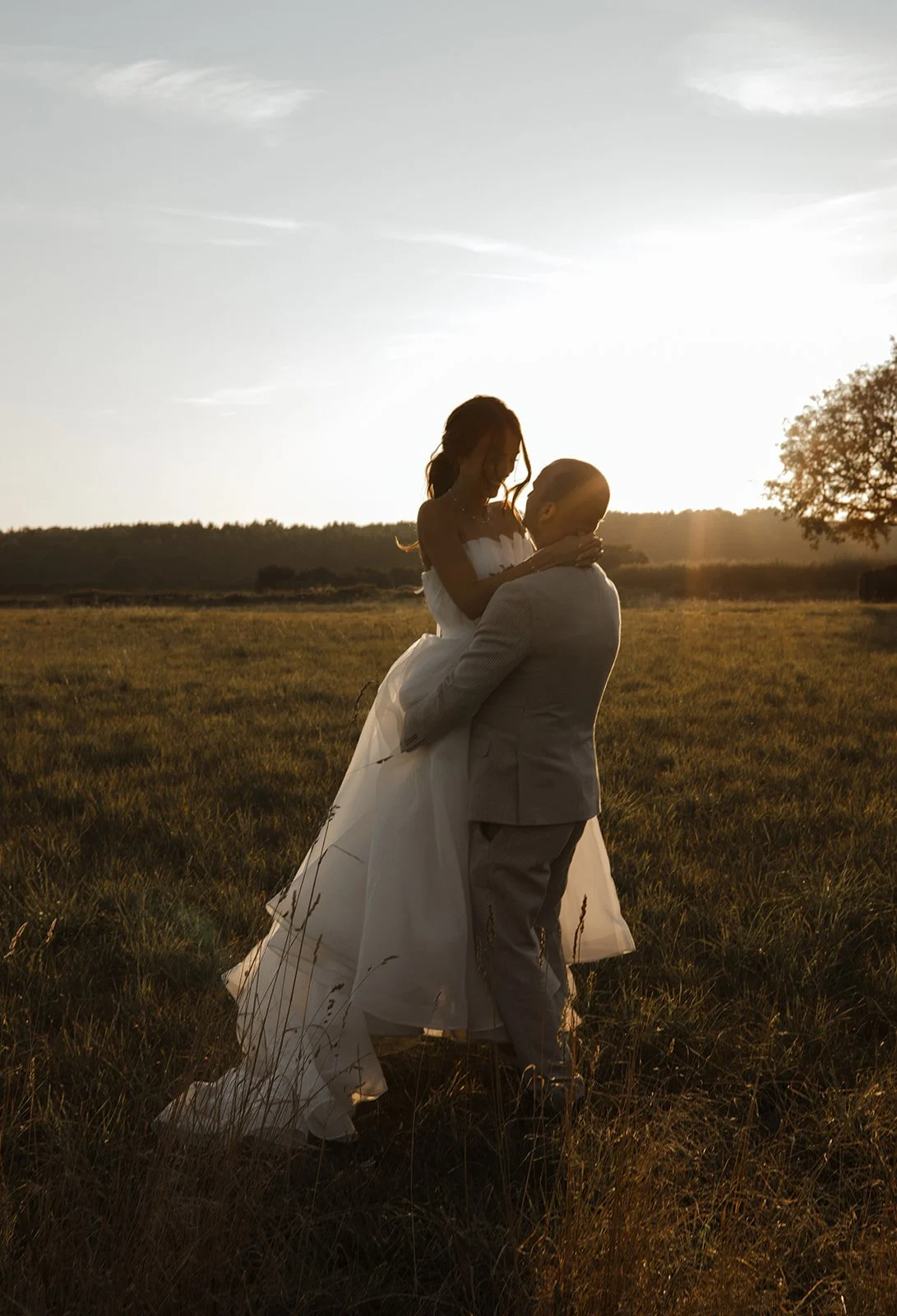 A couple in wedding attire sharing a kiss in a field during sunset, with the bride in a white gown and the groom in a light-colored suit.