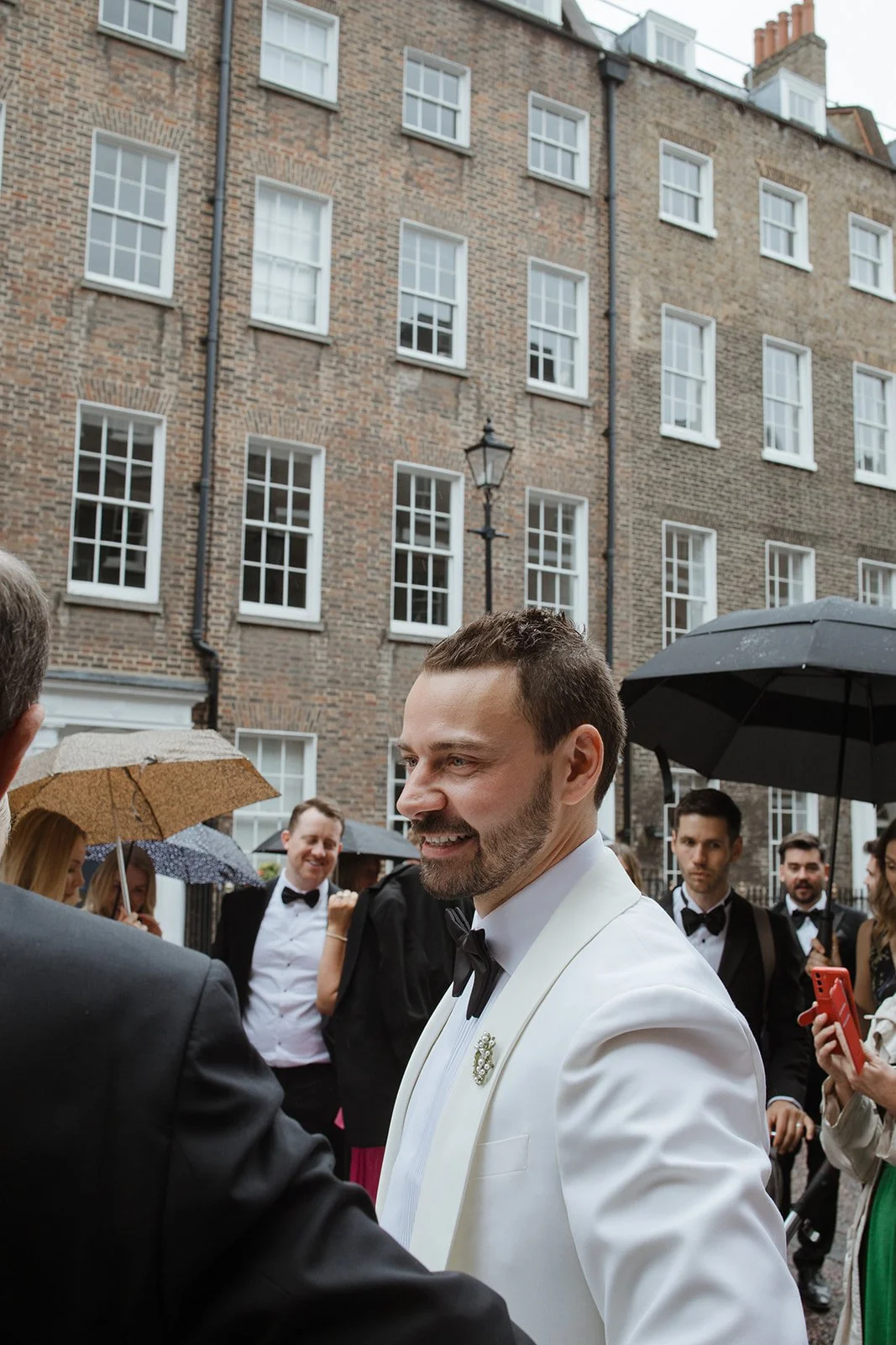 A man in a white tuxedo with a black bow tie smiling at a wedding event outdoors, with a brick building in the background, and other guests in formal attire holding umbrellas.