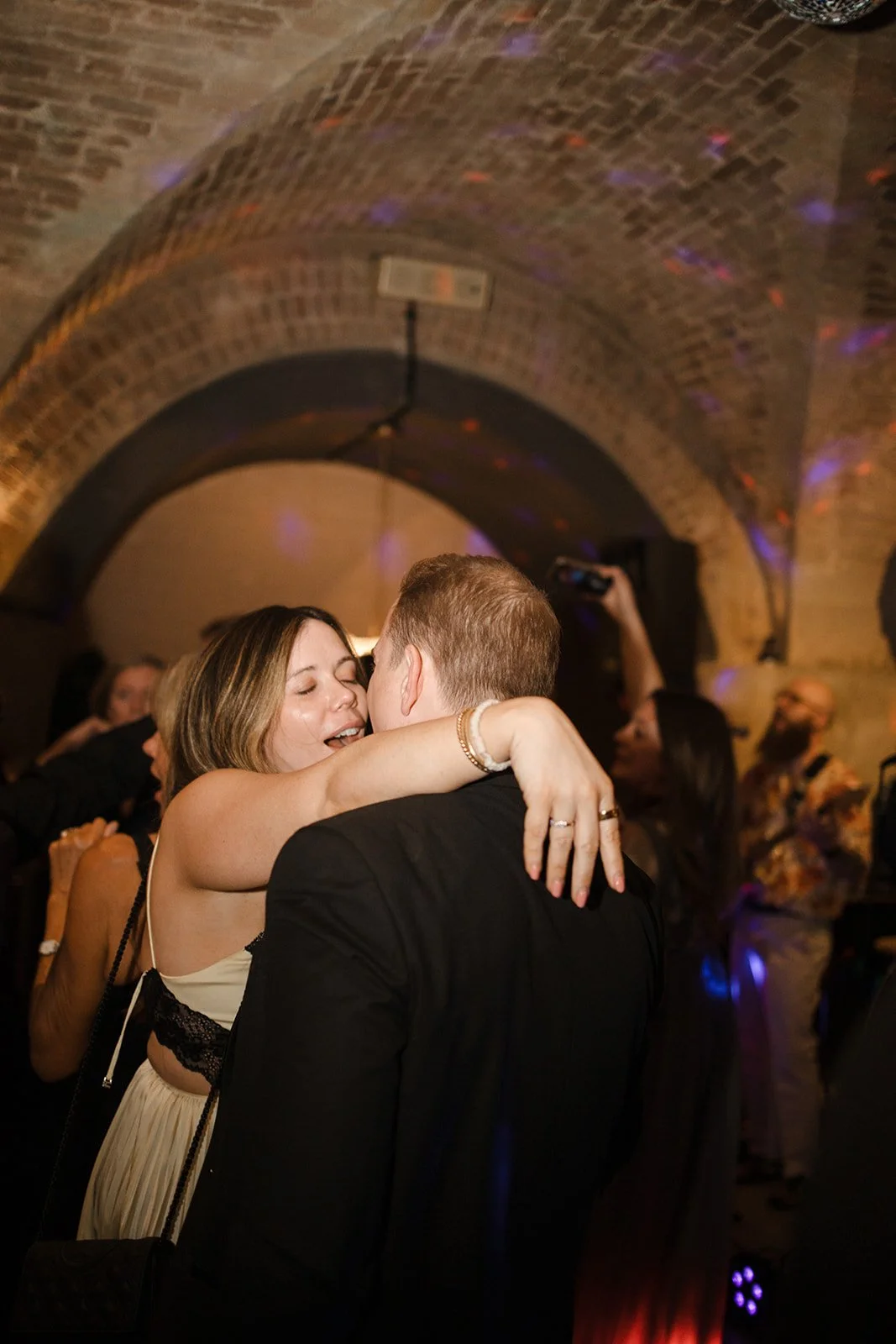 A couple dancing closely at a celebration or wedding with a brick arch ceiling, surrounded by other people enjoying the event.