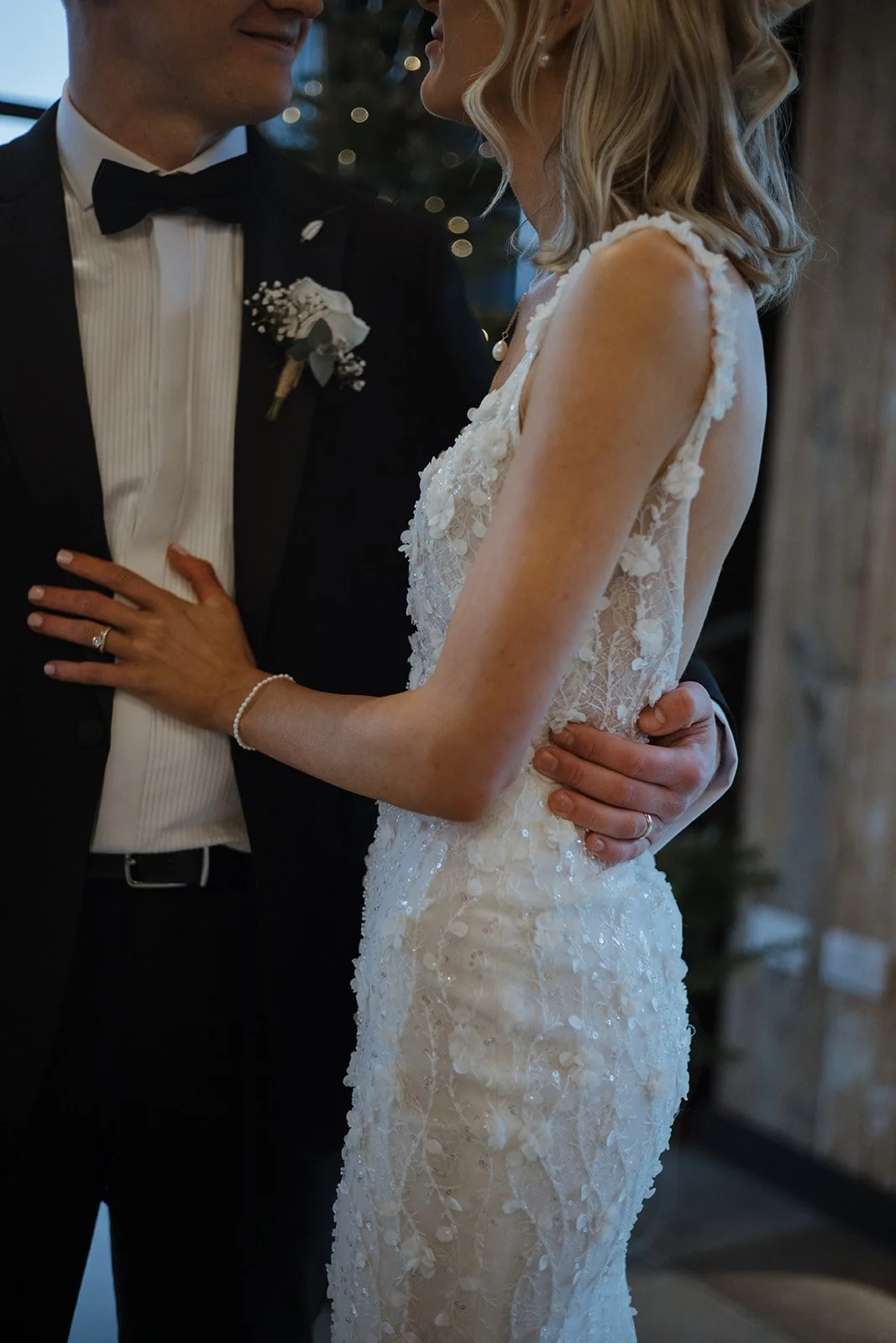 A bride and groom close together, wedding rings visible, bride in a lace gown with floral details, groom in a tuxedo with a boutonniere, holding each other during a wedding ceremony or celebration.
