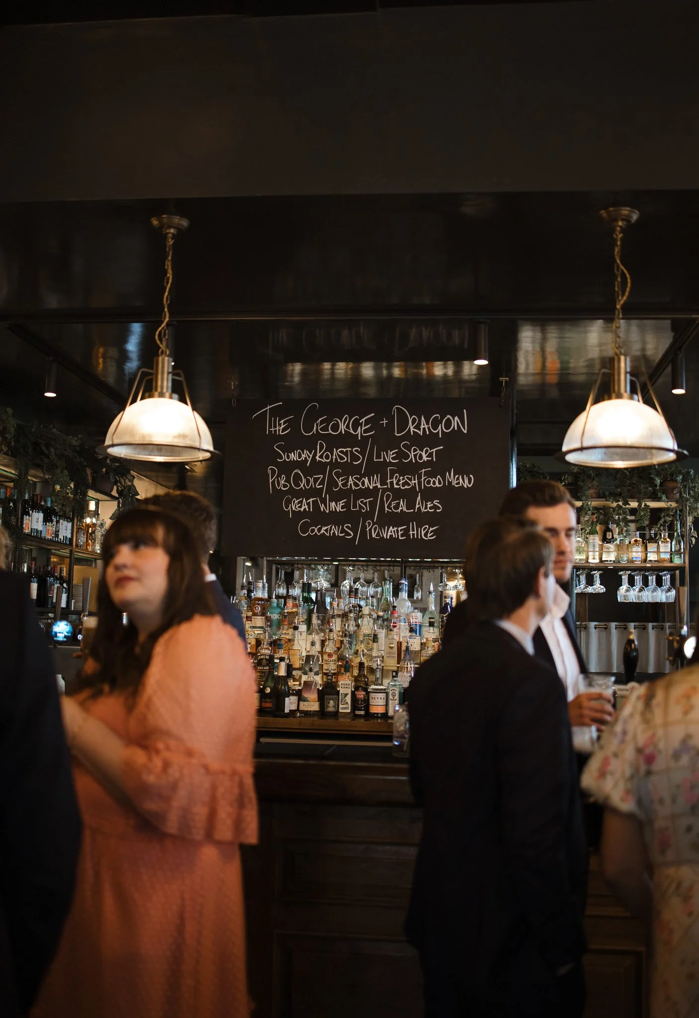 People socializing at a bar in a pub. A chalkboard menu and bottles of liquor are visible behind the bar.