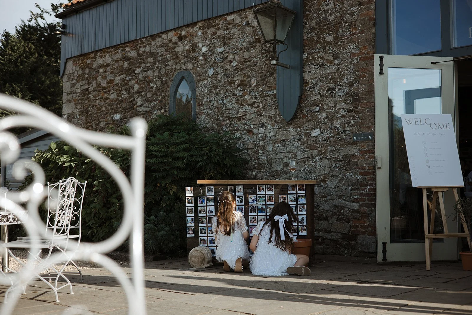 Two girls in white dresses kneeling on the ground, looking at a photo display board outside a building with a brick wall and a sign welcoming guests.