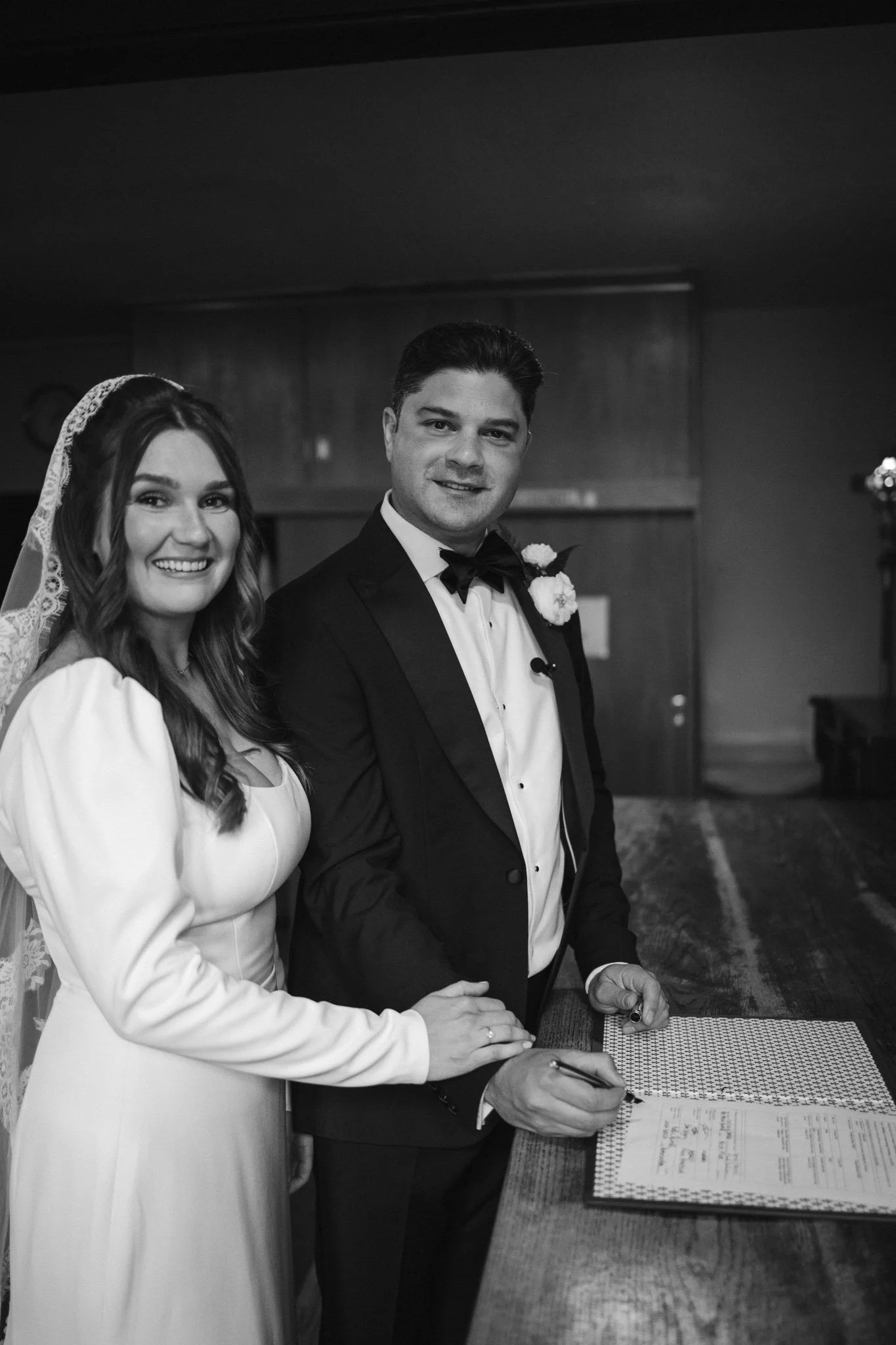 Black and white photo of a bride and groom at their wedding, signing marriage documents at a wooden table.