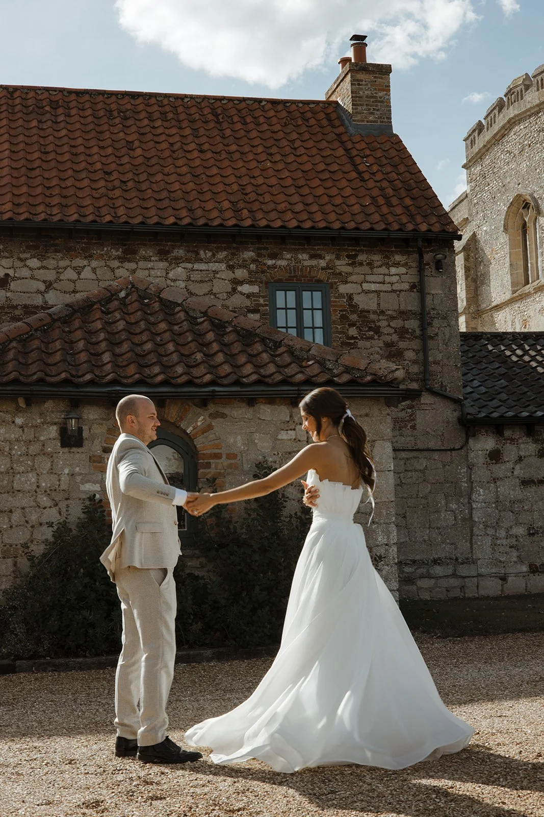 A bride and groom holding hands and dancing outdoors on a gravel surface in front of a stone building with a red tiled roof. The groom wears a light-colored suit, and the bride wears a white wedding dress.