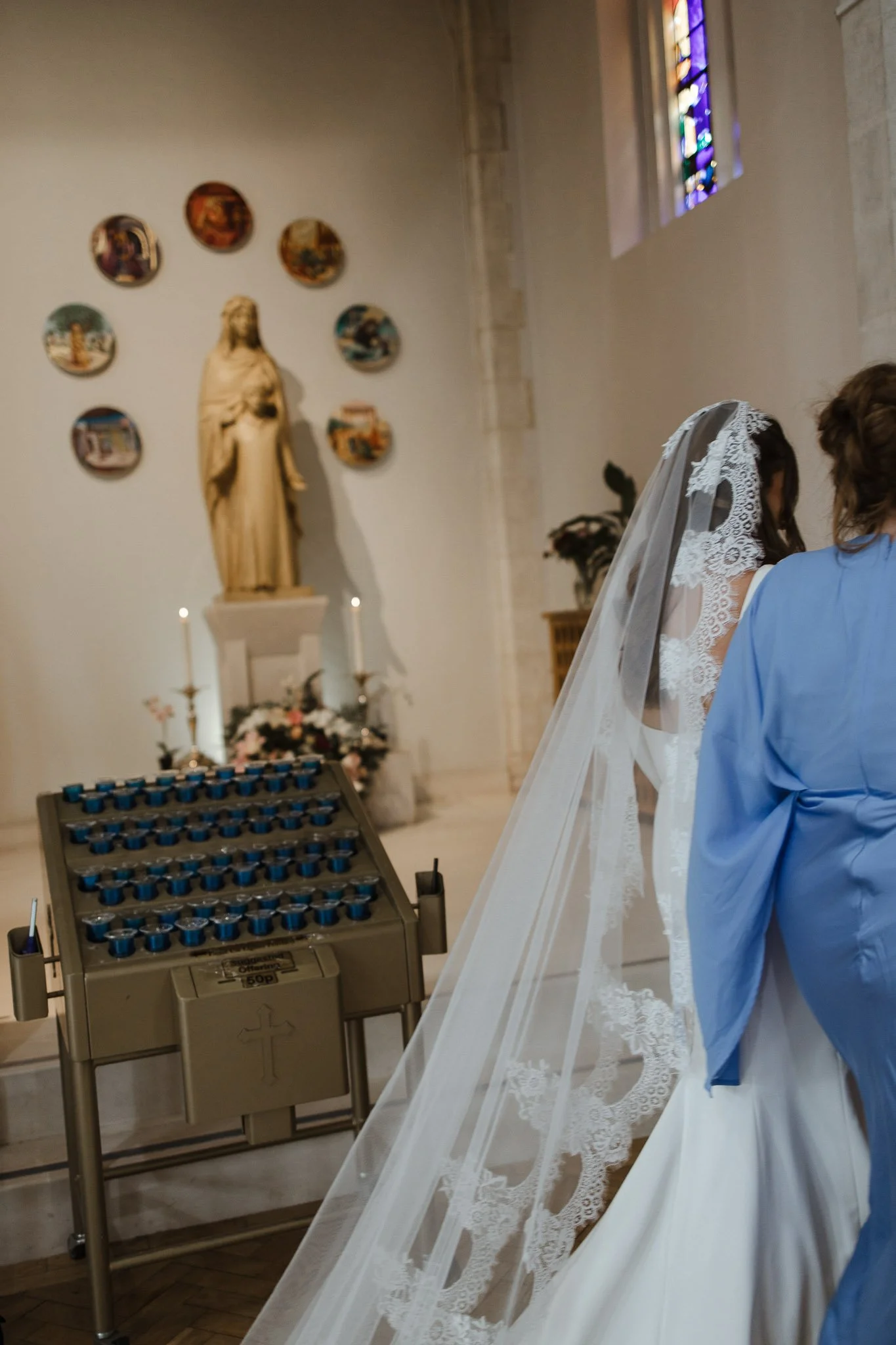 A bride in a white wedding dress with a lace veil, standing next to a woman in a blue robe inside a church. In the background, a statue of Jesus and decorative plates on the wall are visible, along with stained glass windows.